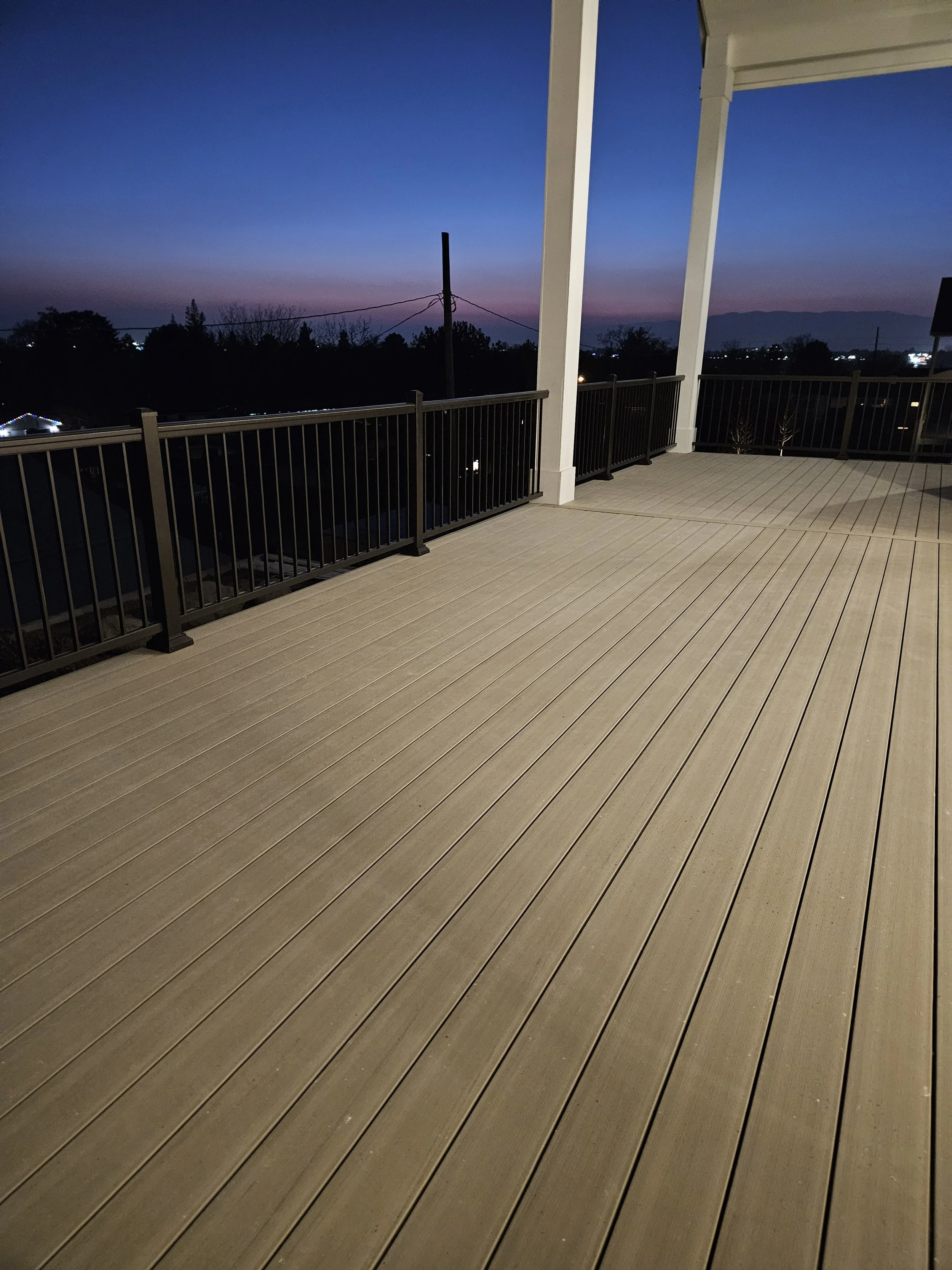 Empty outdoor balcony or deck with beige wood flooring, white columns, black metal railing, and a view of trees and a distant mountain range during dusk or early night.