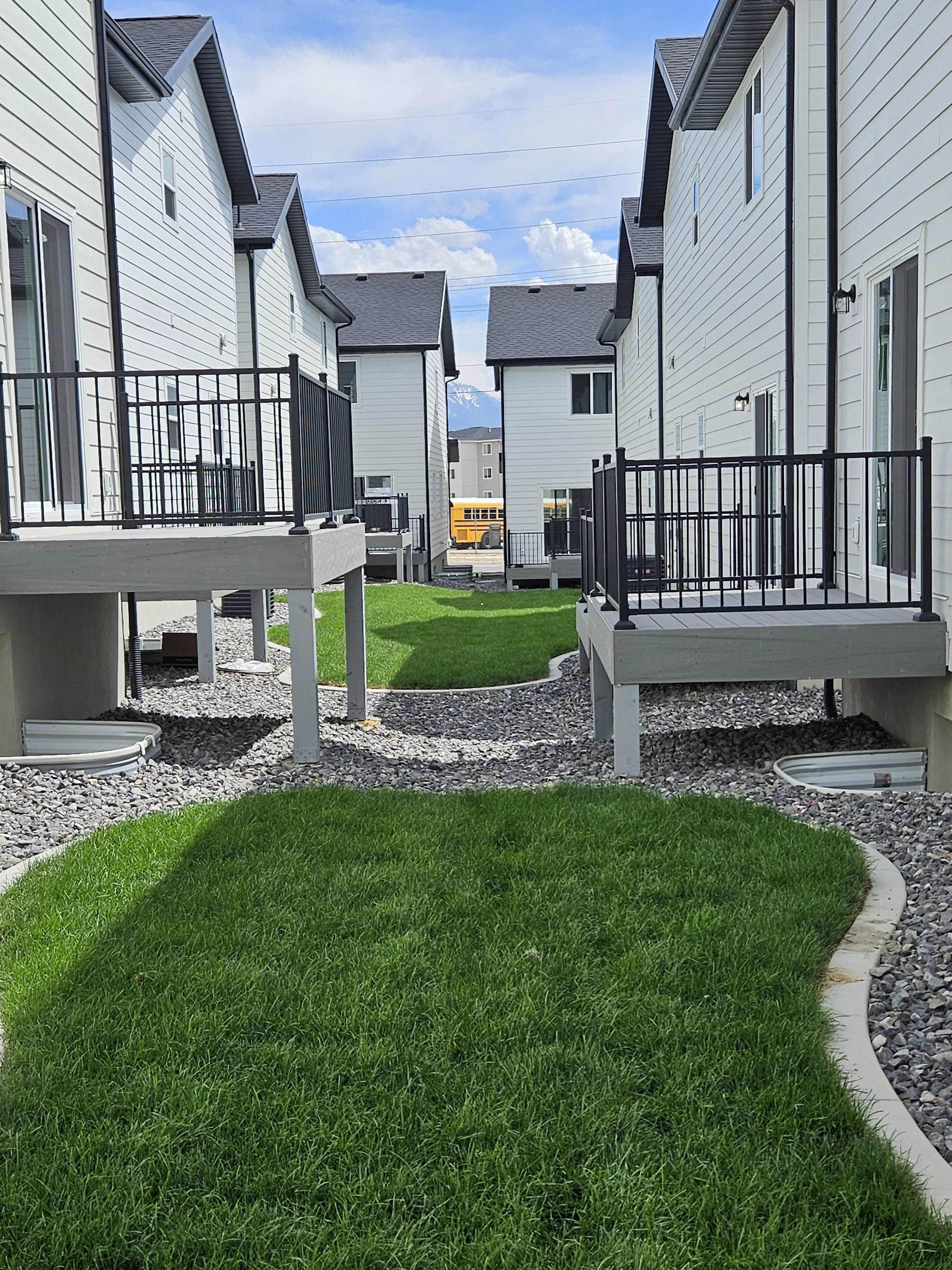 View of a backyard between two white houses with small decks and black railings, a grassy area in the center, and mountains and a school bus in the background.