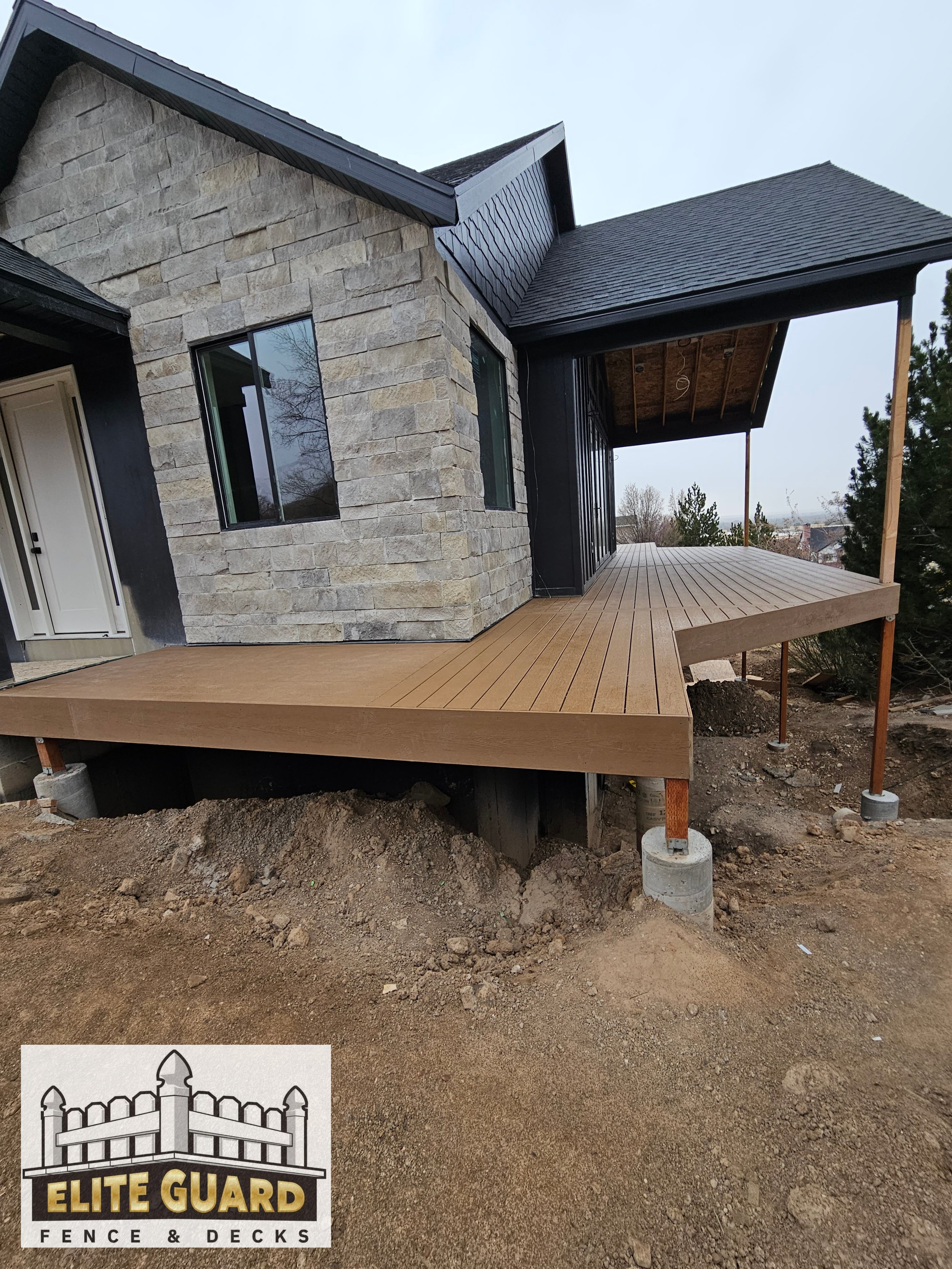A newly constructed house with a stone and black exterior, featuring a wooden deck supported by construction posts and situated on a dirt surface, with trees in the background in Payson, Utah.