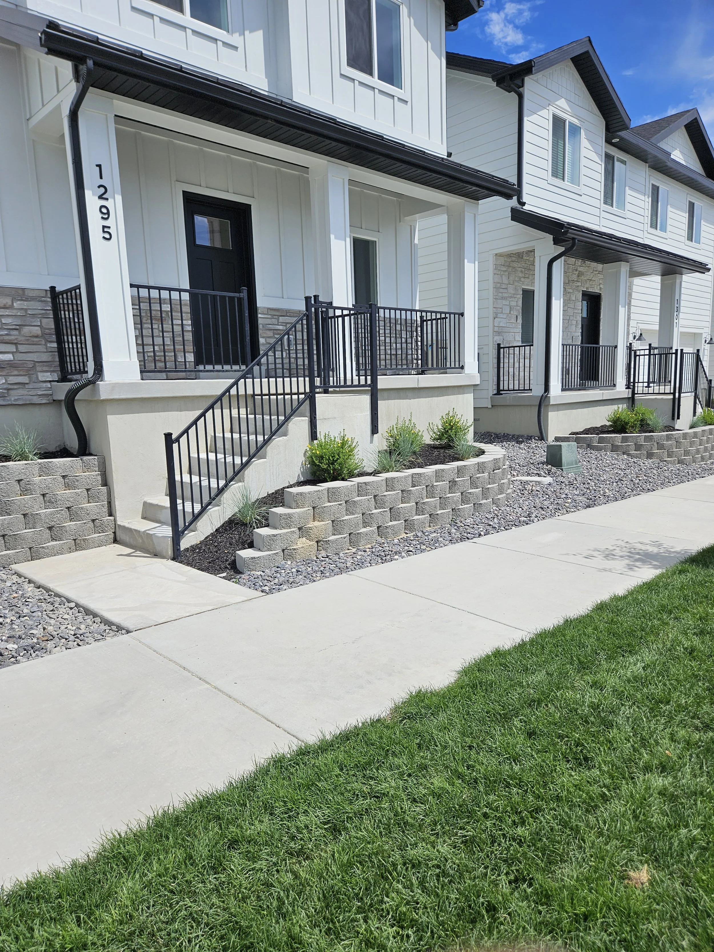 A row of modern townhouses with small front porches, black metal railings, stairs, and landscaped front yards with small bushes, gravel, and paved walkways.