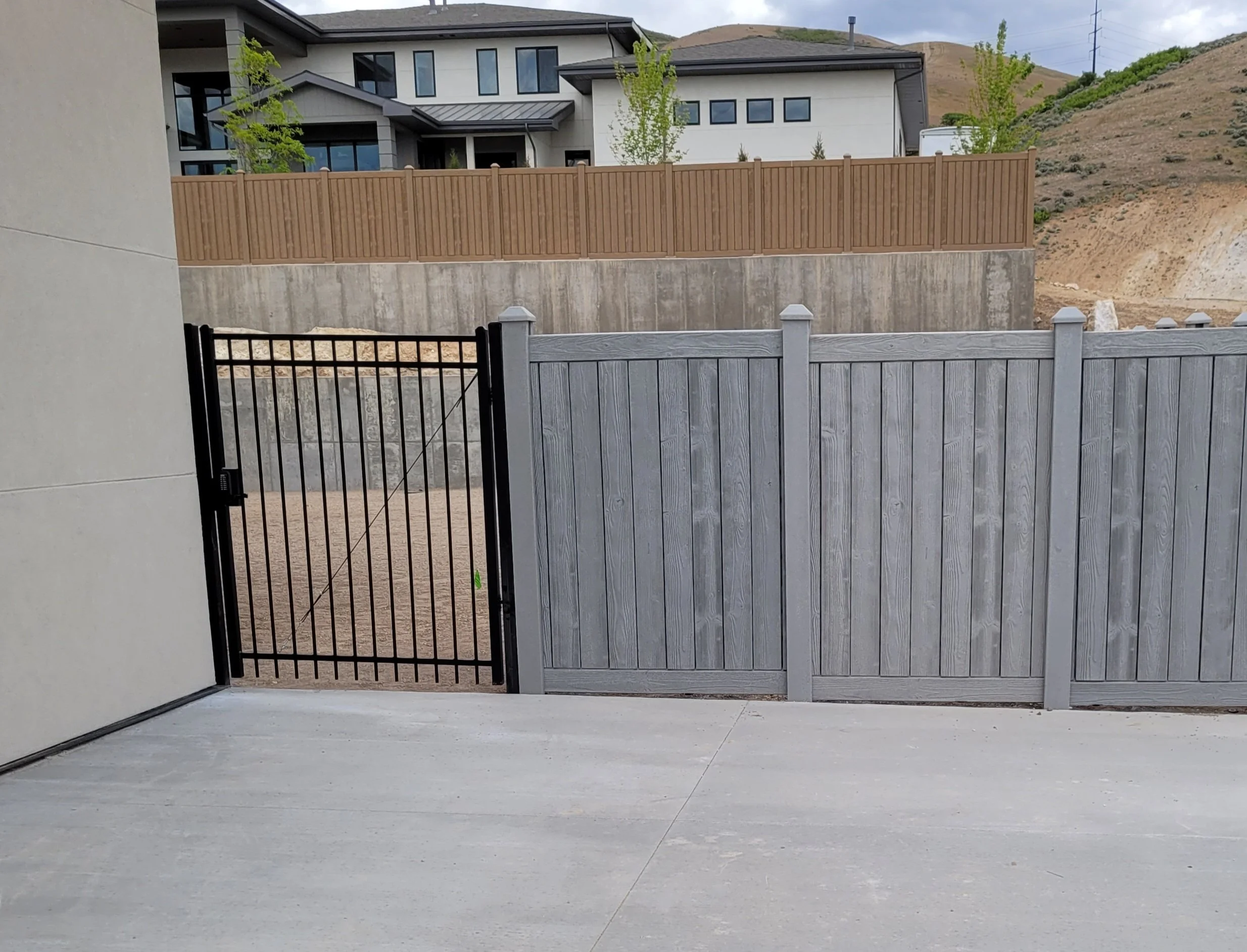 View of a backyard with a black metal gate, wooden fence, and concrete patio in front of a multi-story modern house with large windows, situated on a hillside with dry terrain and hills in the background in Payson, UT.