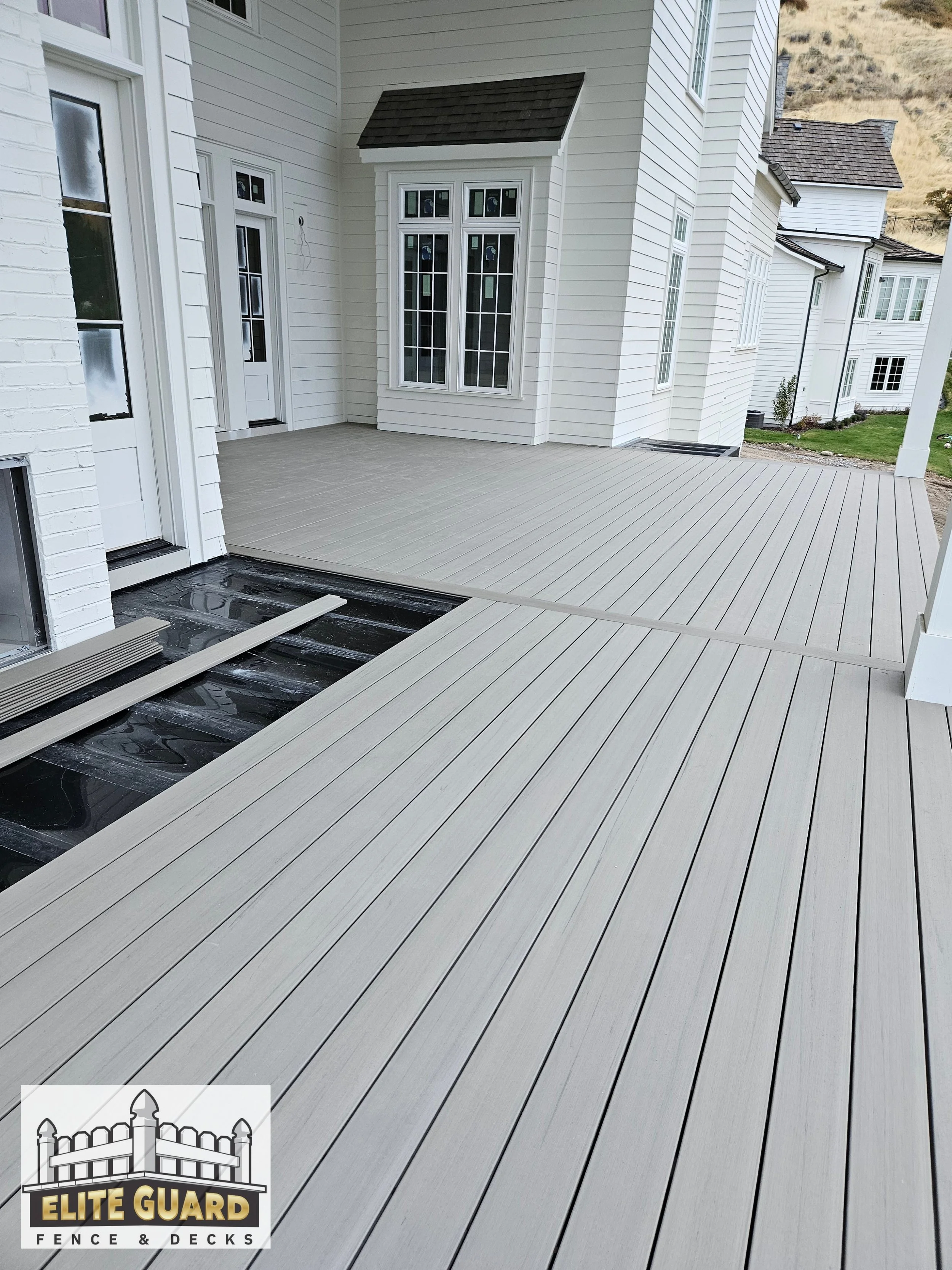 Newly installed gray composite deck on the back porch of a white house, with some construction materials and tools visible, and logo of Elite Guard Fence & Decks in the corner in Spanish Fork, Utah.