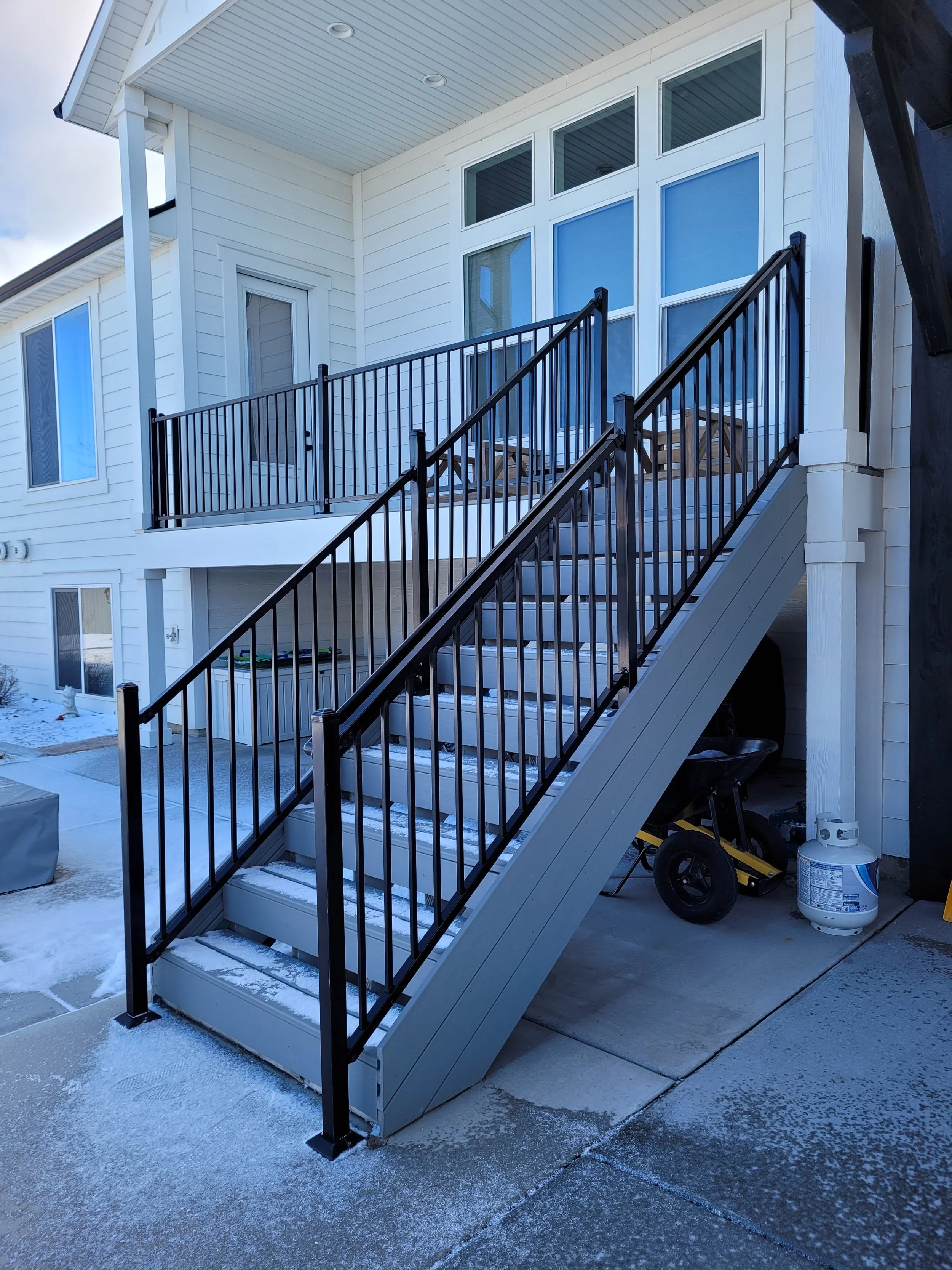 Exterior of a white two-story house with a metal staircase leading to a second-floor balcony. The staircase has black metal railings and snow on the steps. The balcony has white siding and large windows.