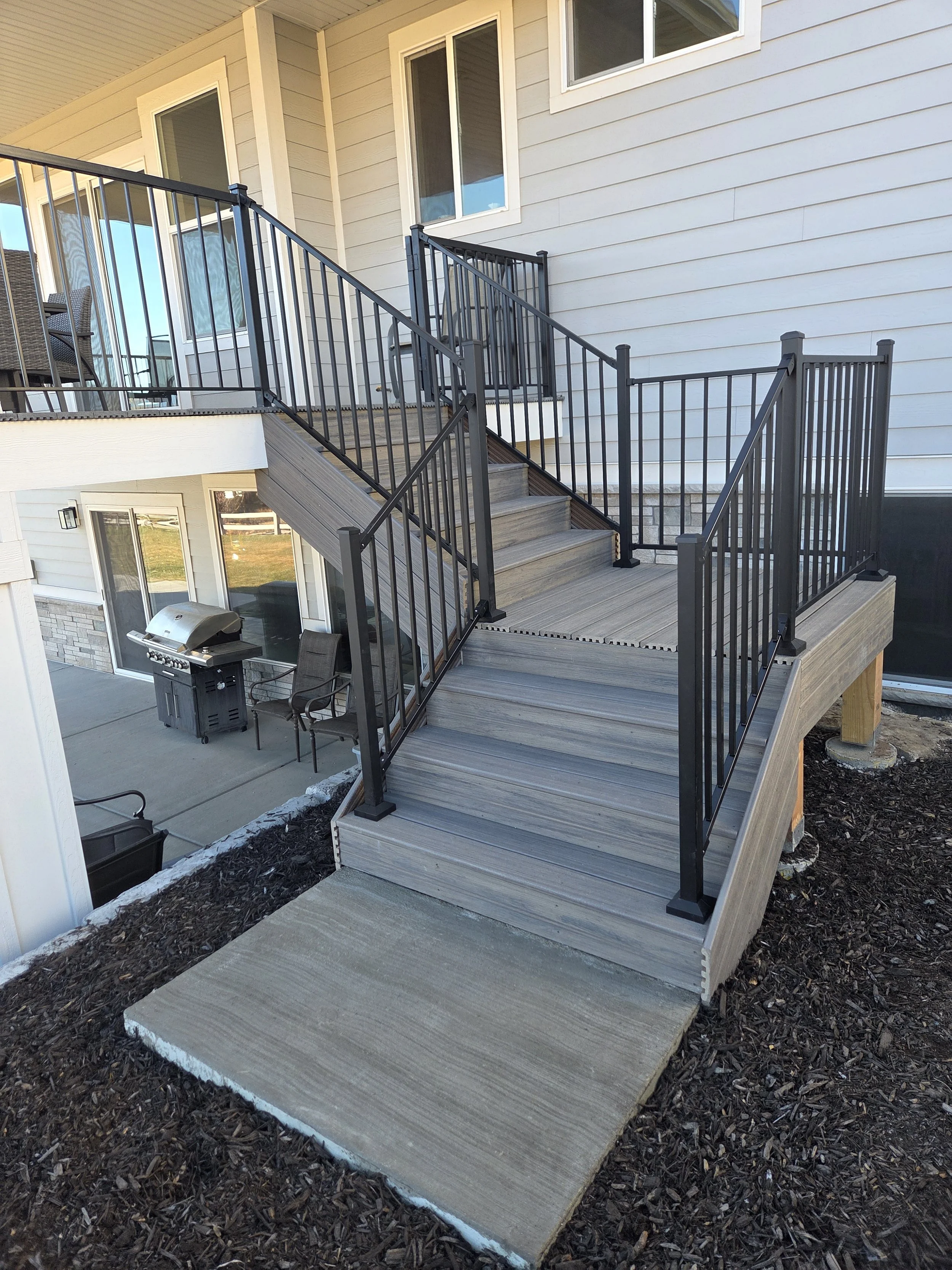 Outdoor wooden staircase with black metal railings leading to a second-floor balcony of a house. The house has beige siding and multiple white-framed windows. A grill and chairs are visible on the lower patio area in Payson, UT.