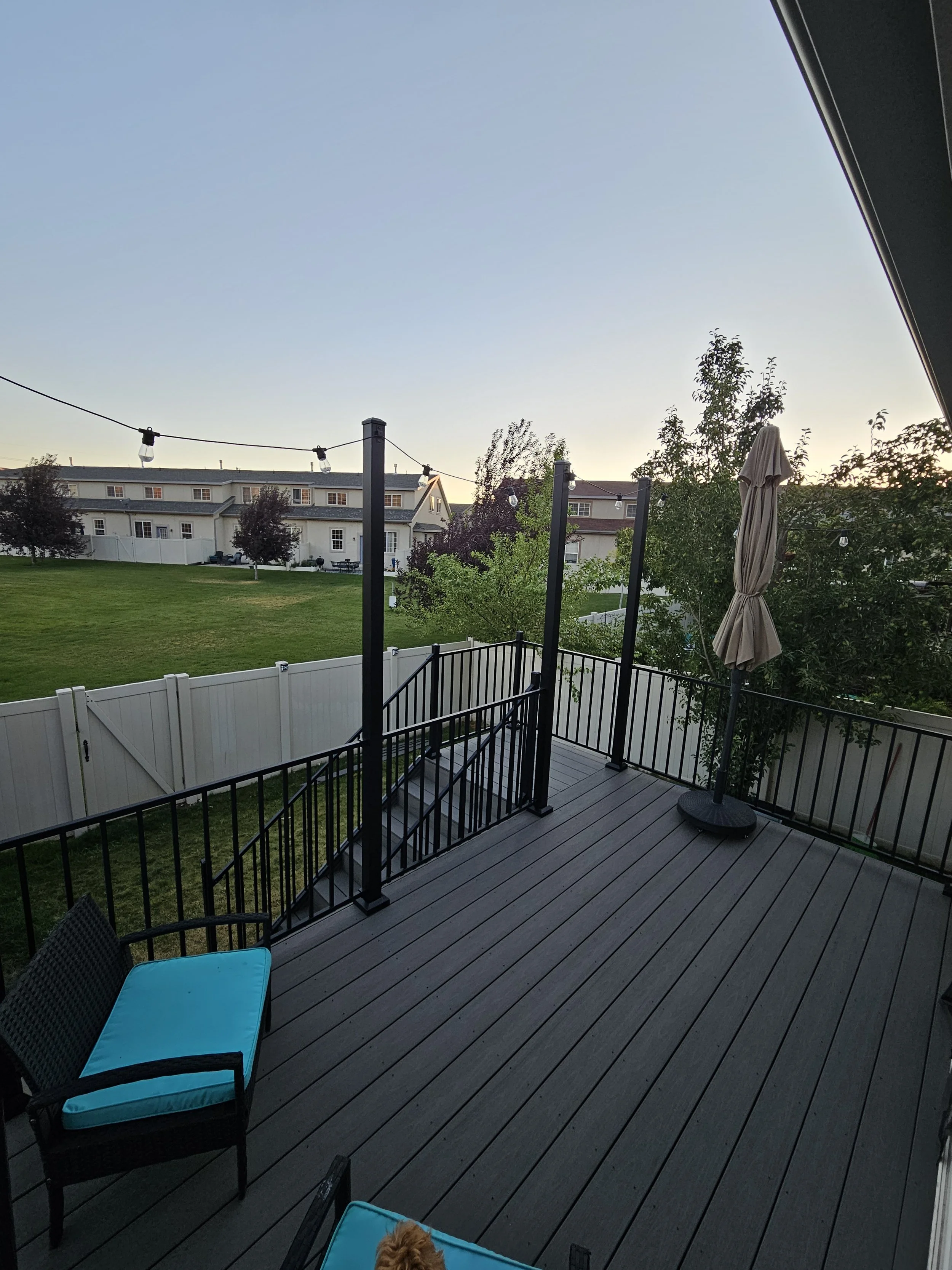A backyard deck with black railings, a closed beige patio umbrella, a chair with a turquoise cushion, and a tree with green leaves, overlooking a grassy area and neighboring houses in the distance during sunset.