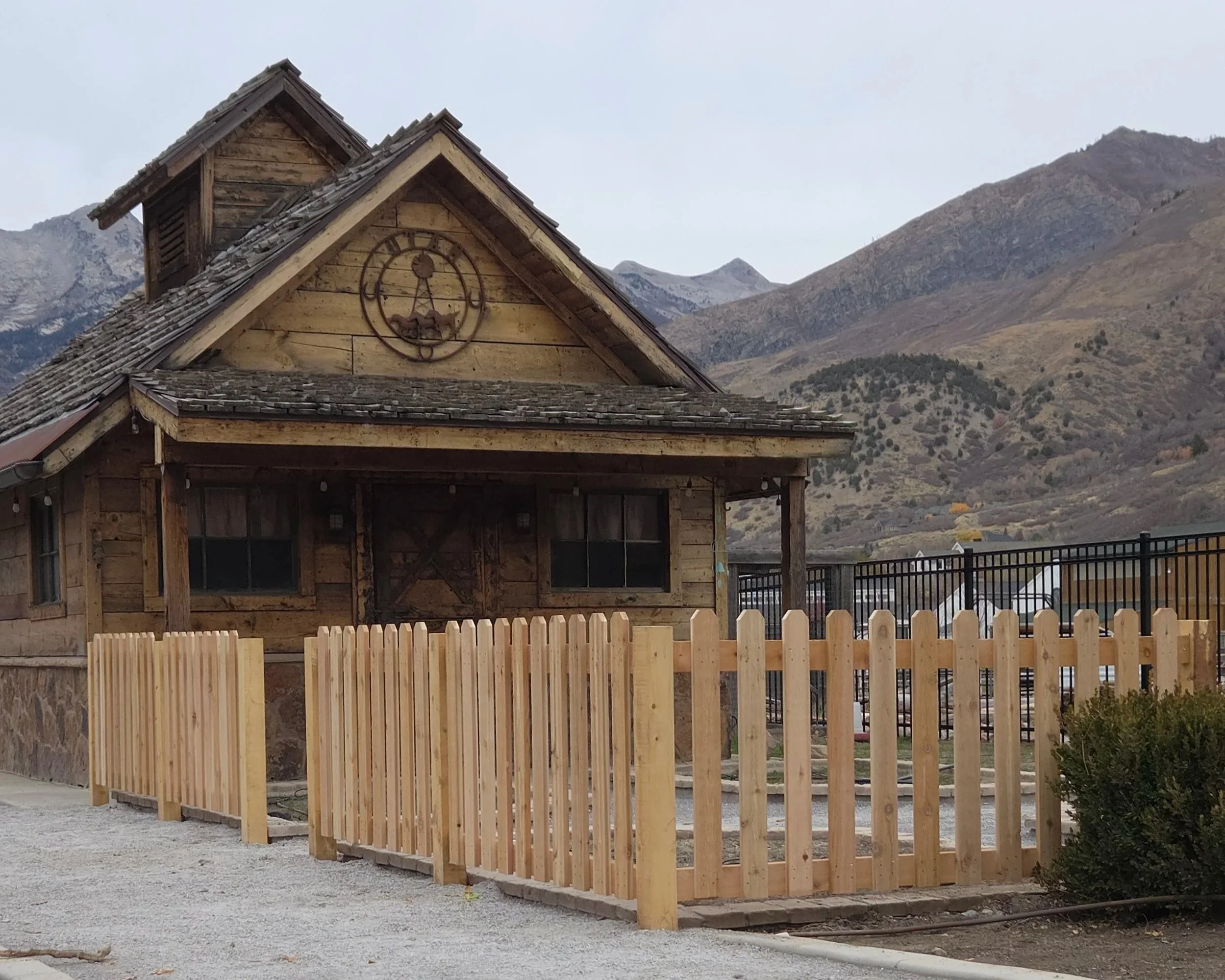 A rustic wooden building with a clock on the front, surrounded by a new wooden fence, with mountains in the background and an overcast sky in Payson, UT.