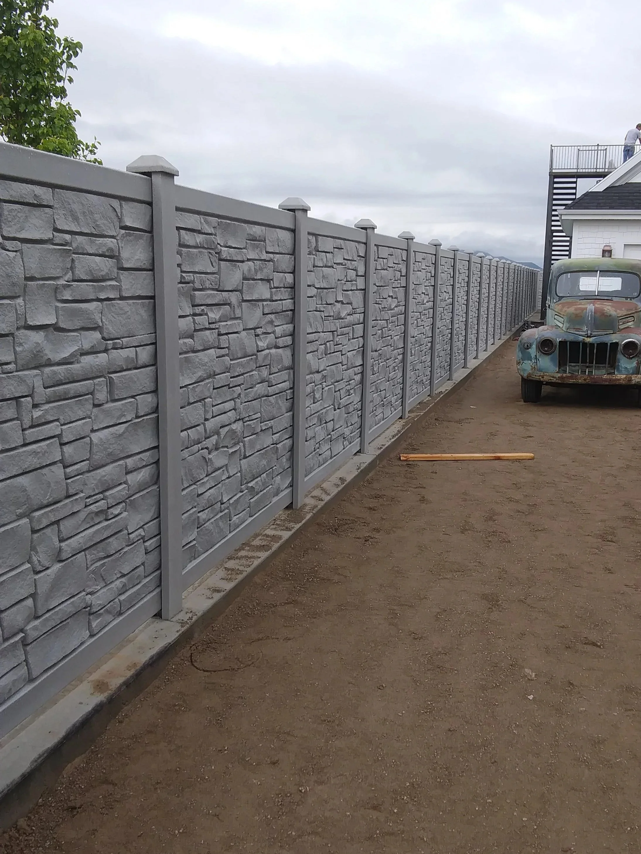 A long grey stone-patterned privacy fence next to a dirt area, with an old rusted truck parked nearby and a house in the background in Springville, UT.
