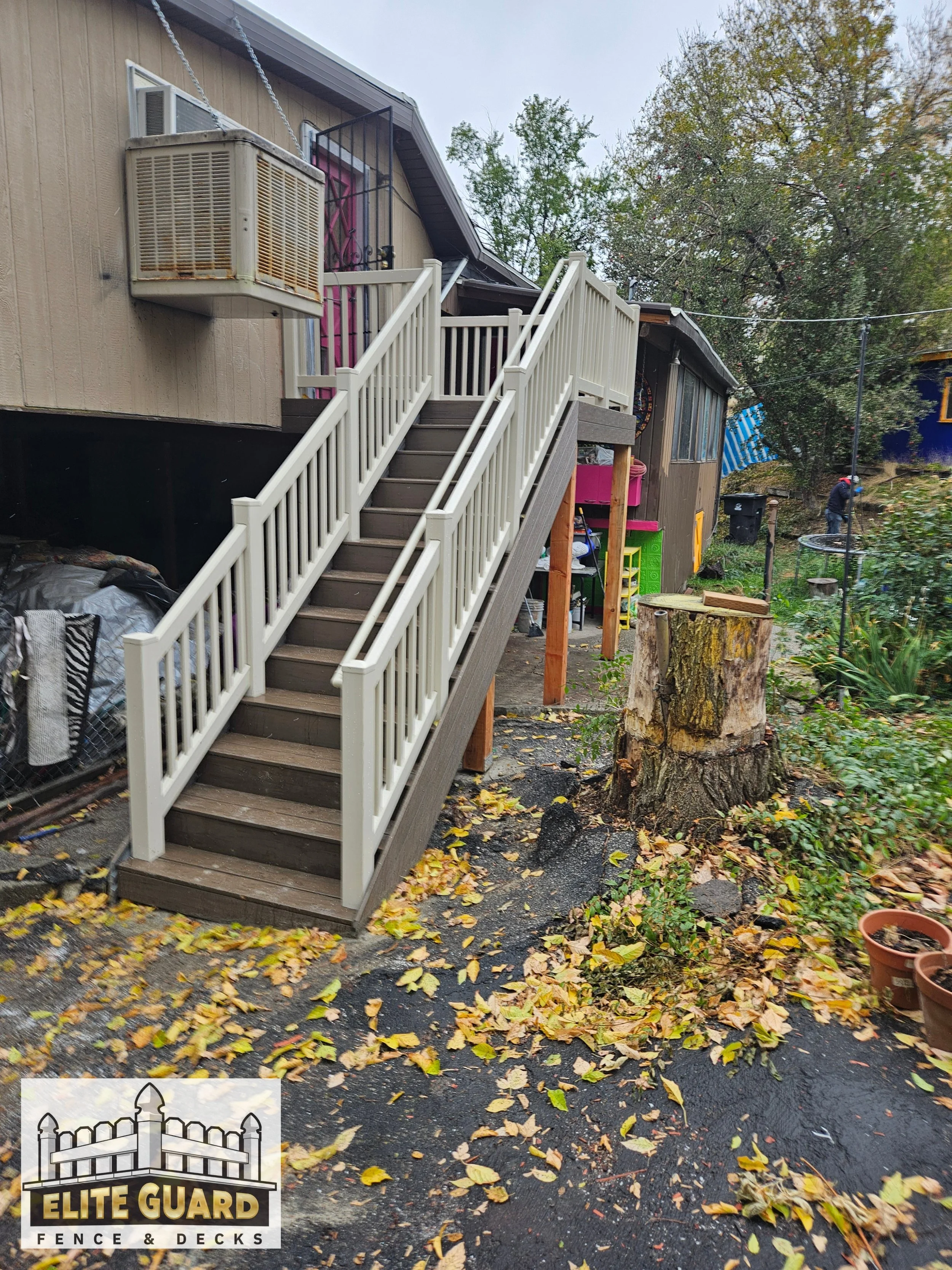 Exterior view of a house with a wooden staircase leading to a deck. The deck has white railings and is elevated. There is a large tree stump in the yard and fallen leaves scattered on the pavement. The yard contains various garden items, plants, and 