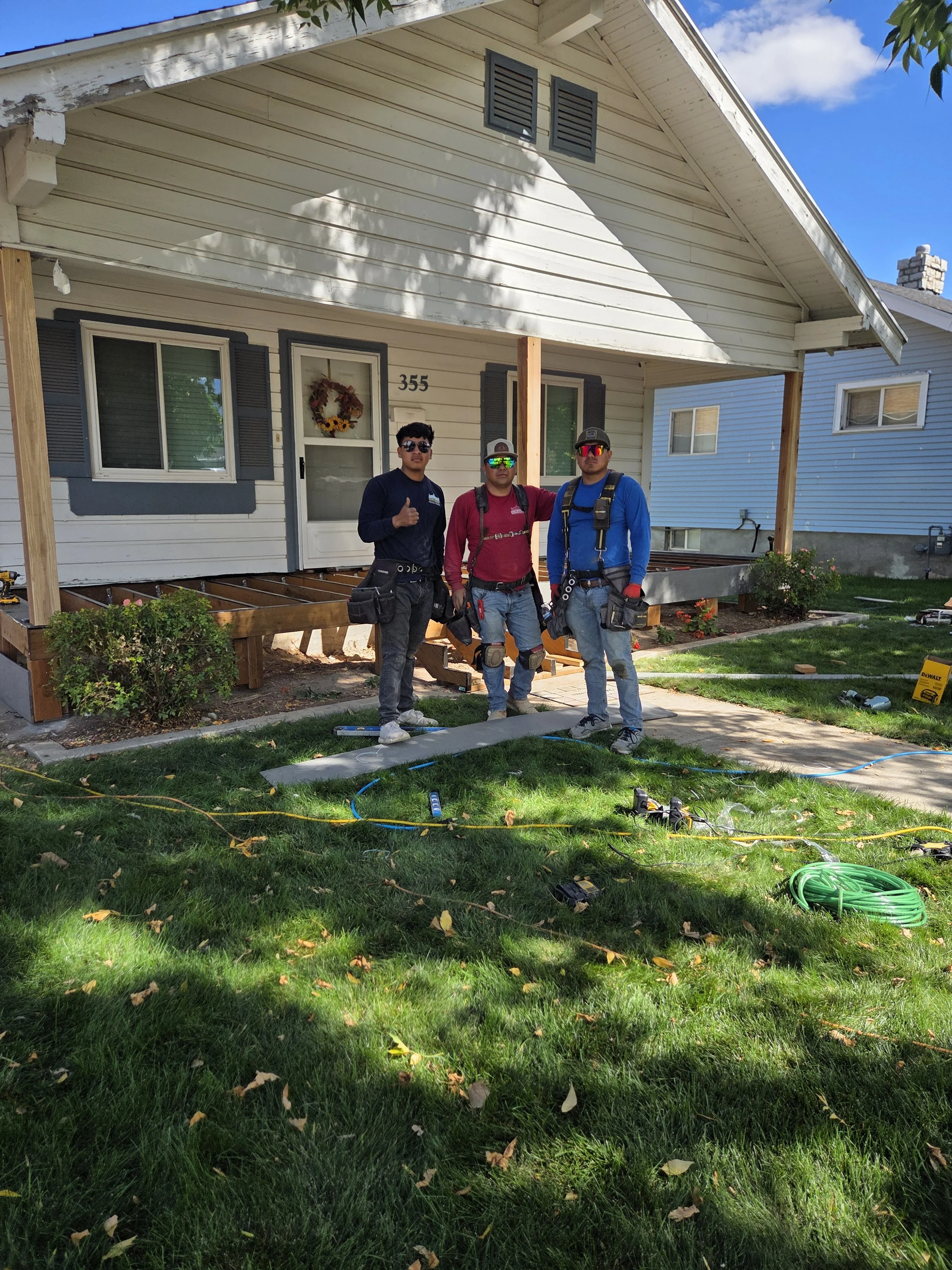 Three workers stand in front of a house under renovation, surrounded by construction tools and electrical cords. The house has a porch with missing steps and a wreath on the door in Payson, UT.