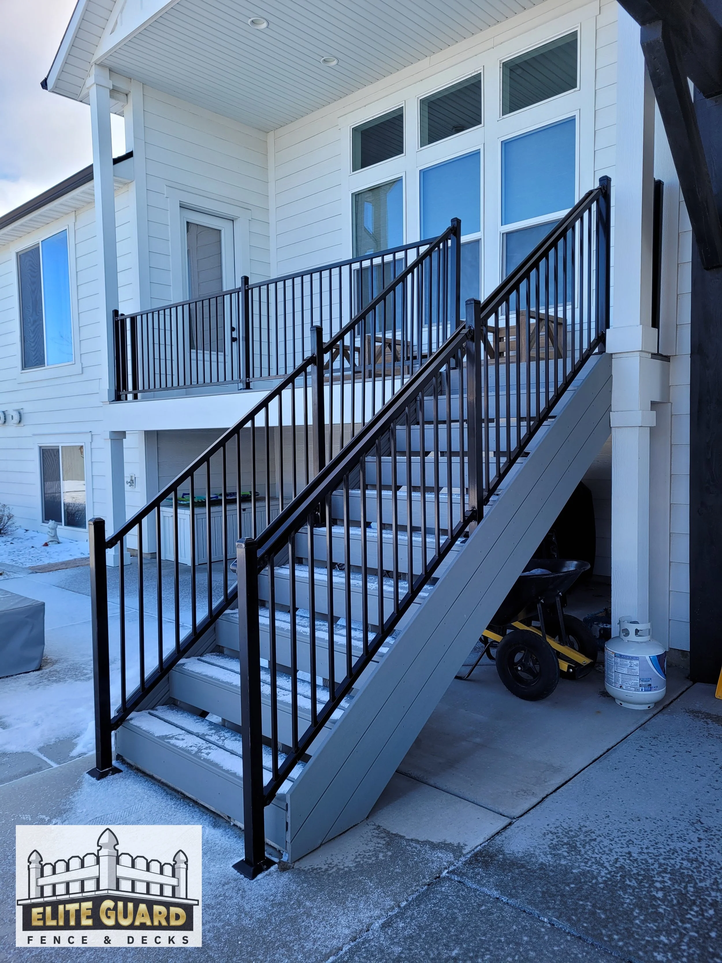 Outdoor staircase with black metal railing leading up to a second-story balcony of a white house with large windows, snow on the ground, and tools and propane tank underneath in Payson, Utah.