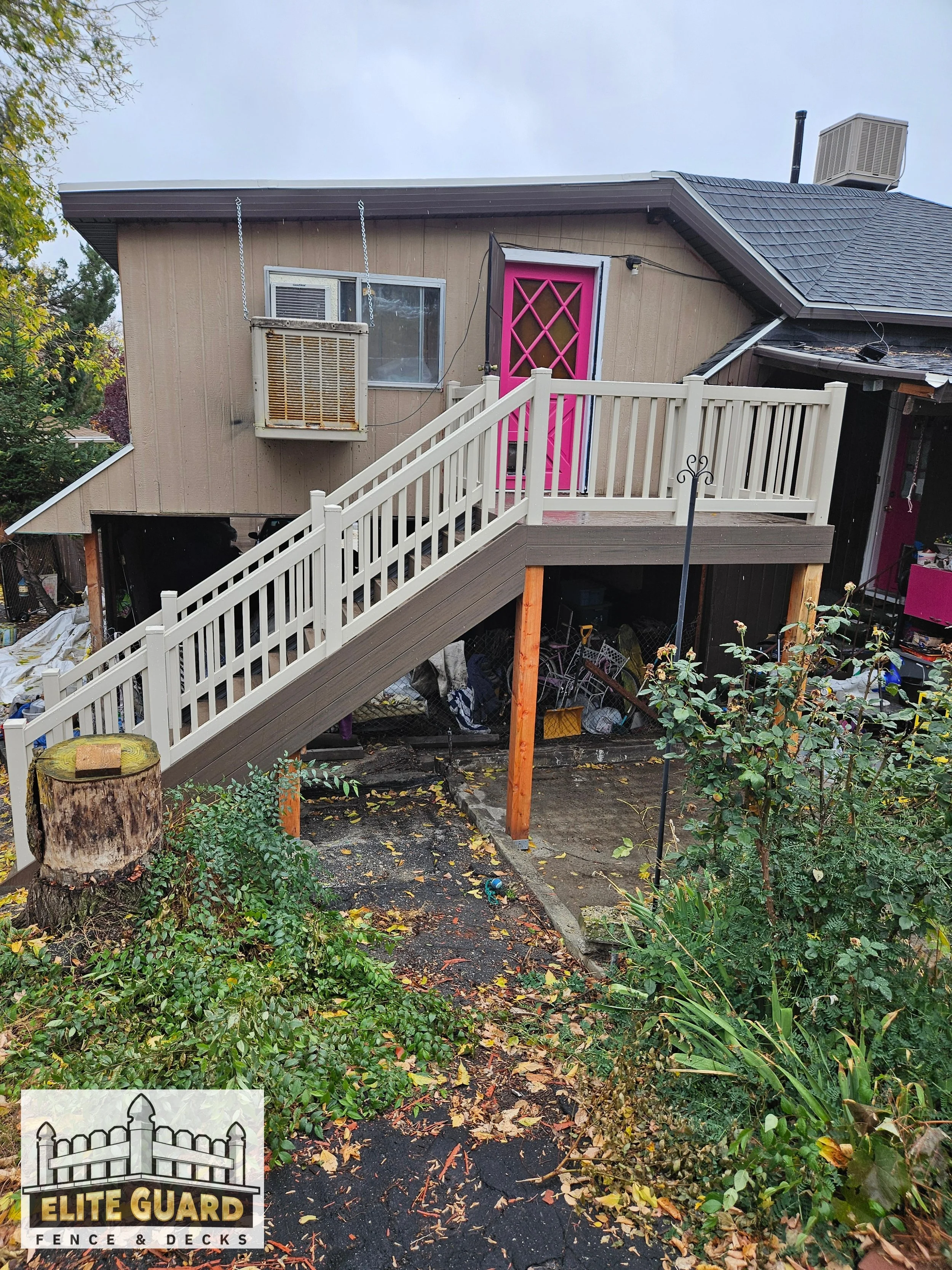 Backyard with stairs leading to an elevated deck. The deck has a pink door, white railing, and beige siding. There is a tree stump, plants, and fallen leaves on the ground in Spanish Fork, Utah.