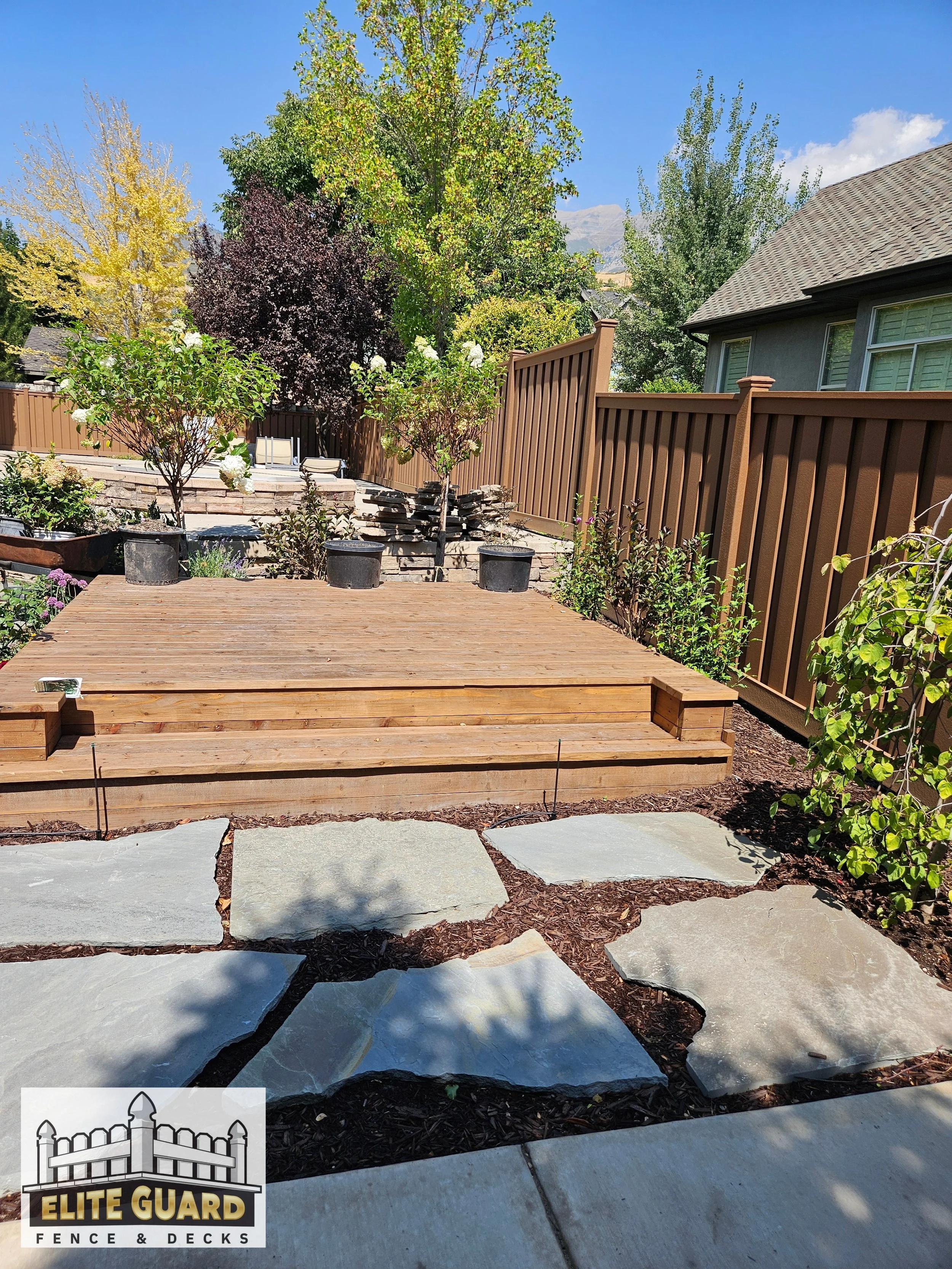 Backyard garden with a wooden deck, large flat stones in a pathway, potted plants on the deck, a brown fence, trees with green and purple leaves, a house with gray siding, and clear blue sky in Spanish Fork, Utah.