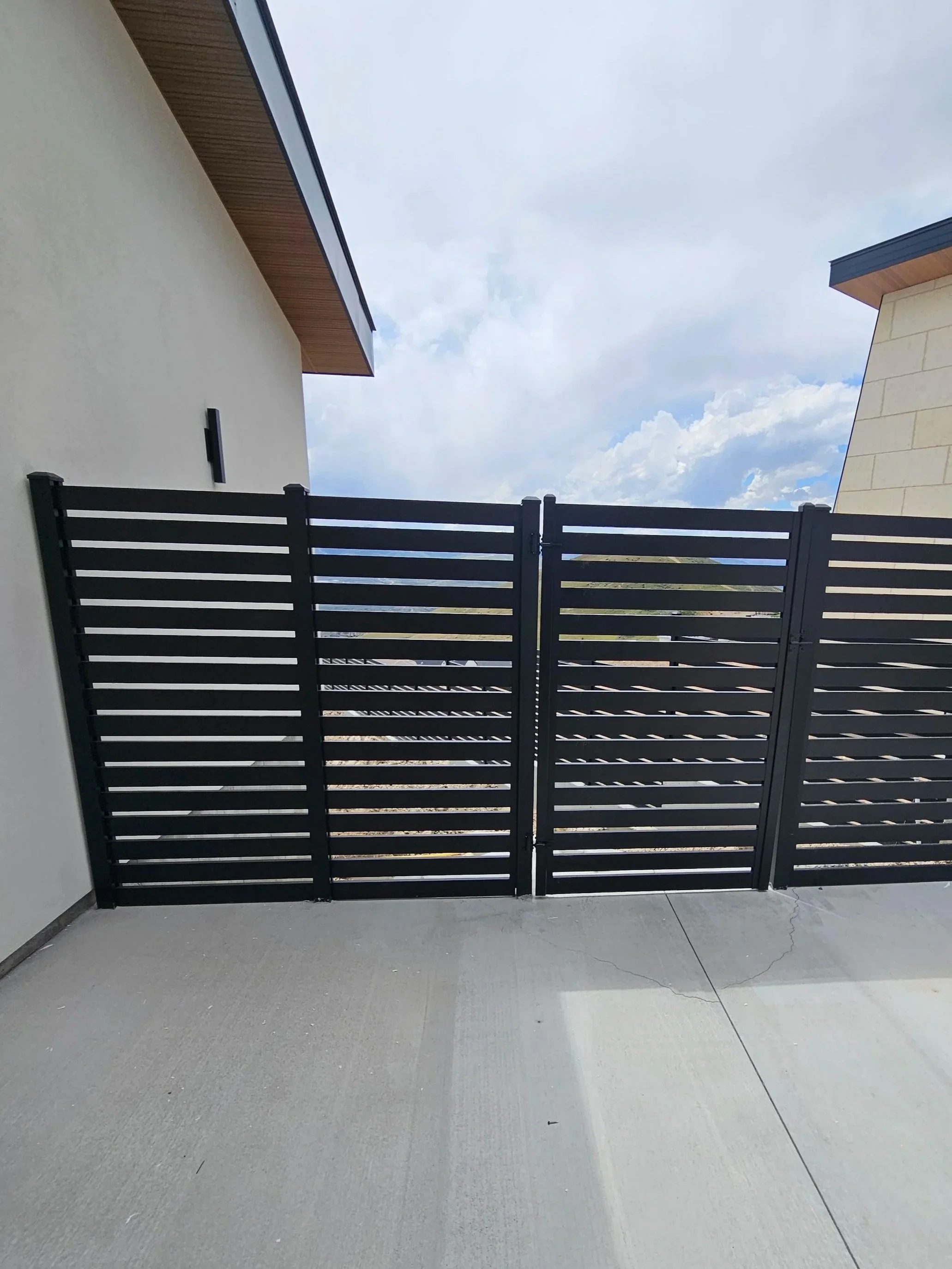 Black horizontal slat privacy fence on a concrete patio with white exterior walls and a partly cloudy sky in Payson, UT.