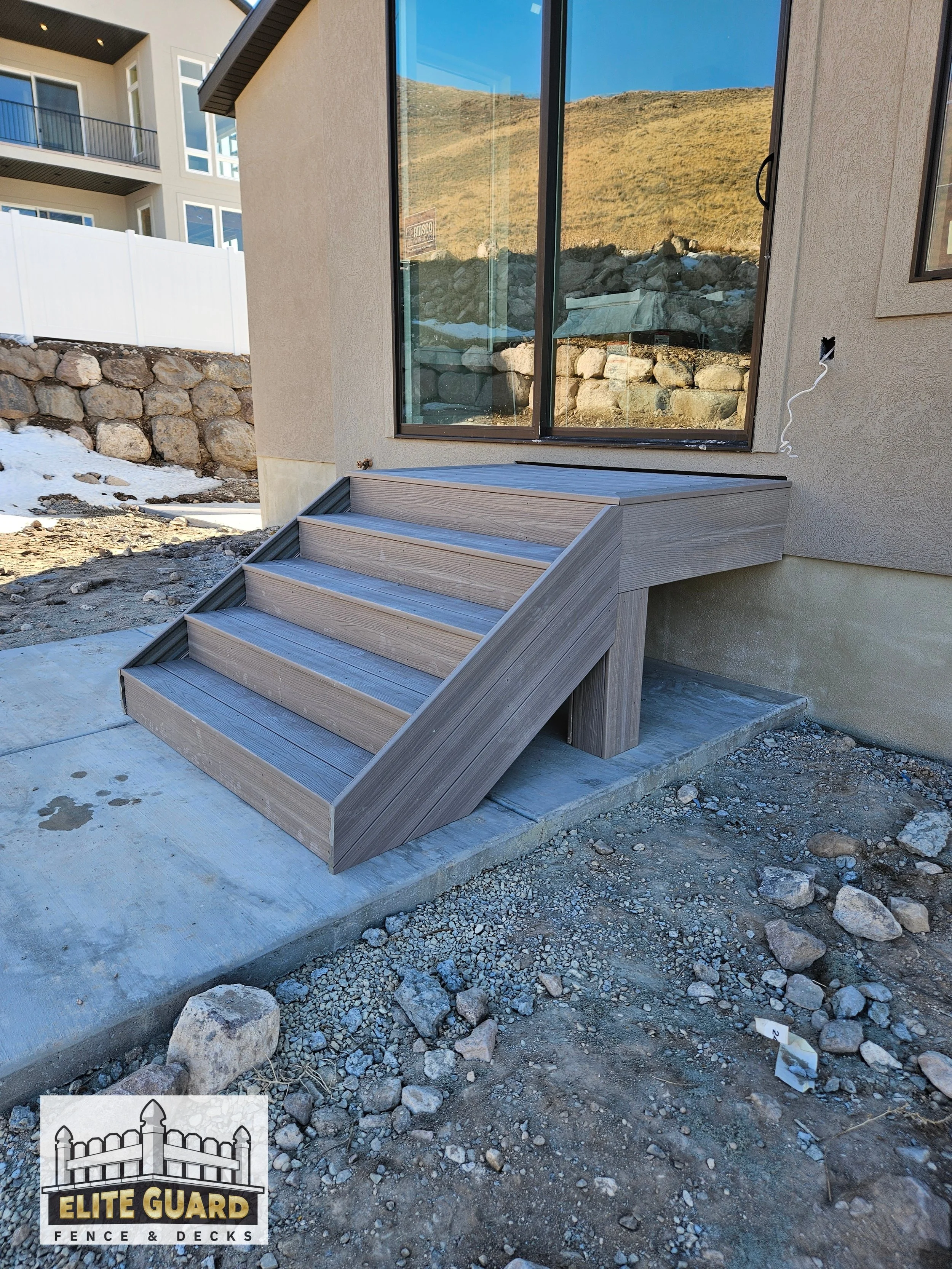 A small wooden staircase with four steps leading up to a patio area outside a house with a large glass door. The house has beige exterior walls. The ground around the stairs is unfinished with rocks and dirt. A logo in the bottom left corner reads 'E