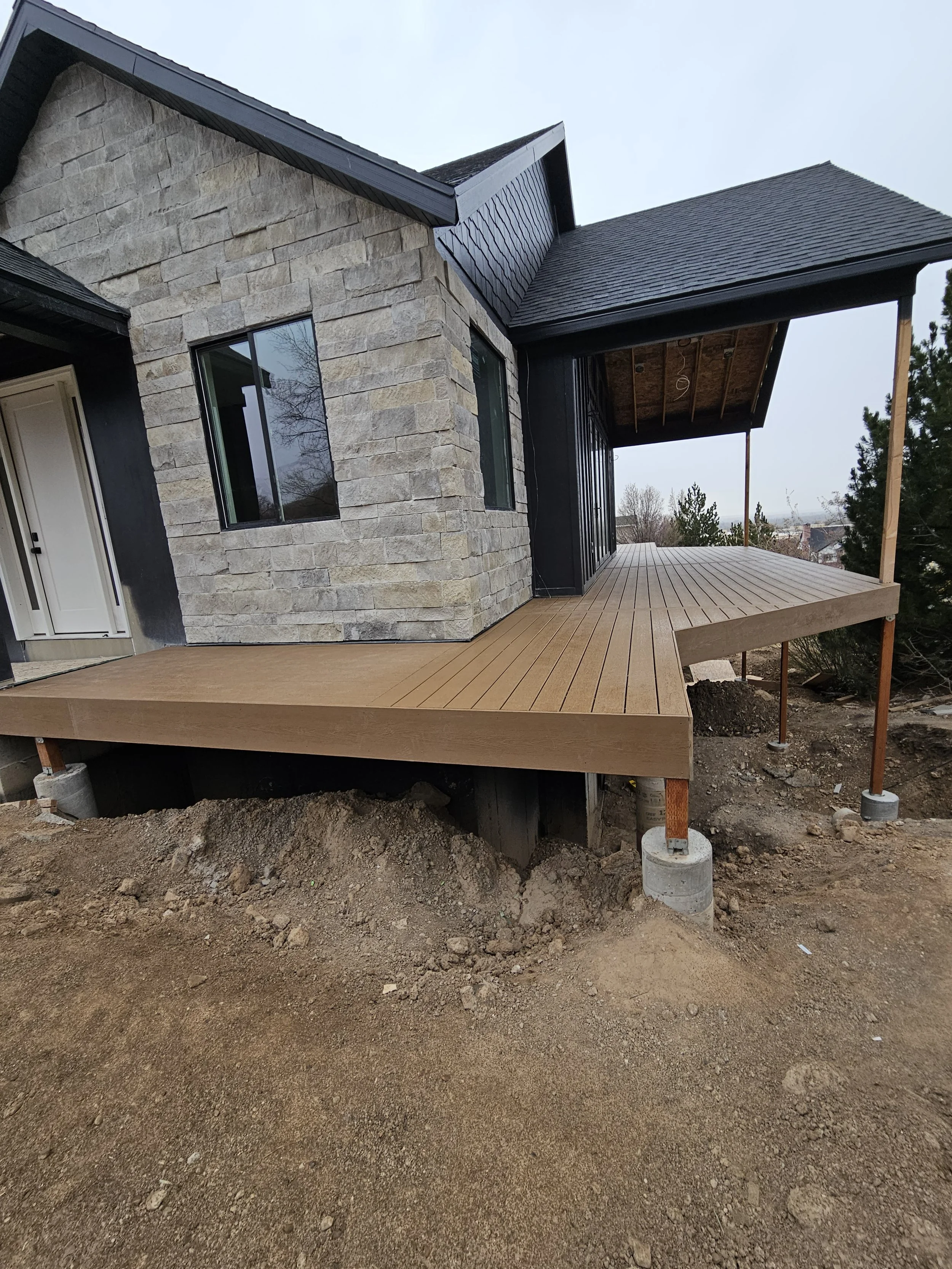 Newly constructed house with a wooden deck, stone and black siding exterior, and multiple windows, located on a dirt lot.