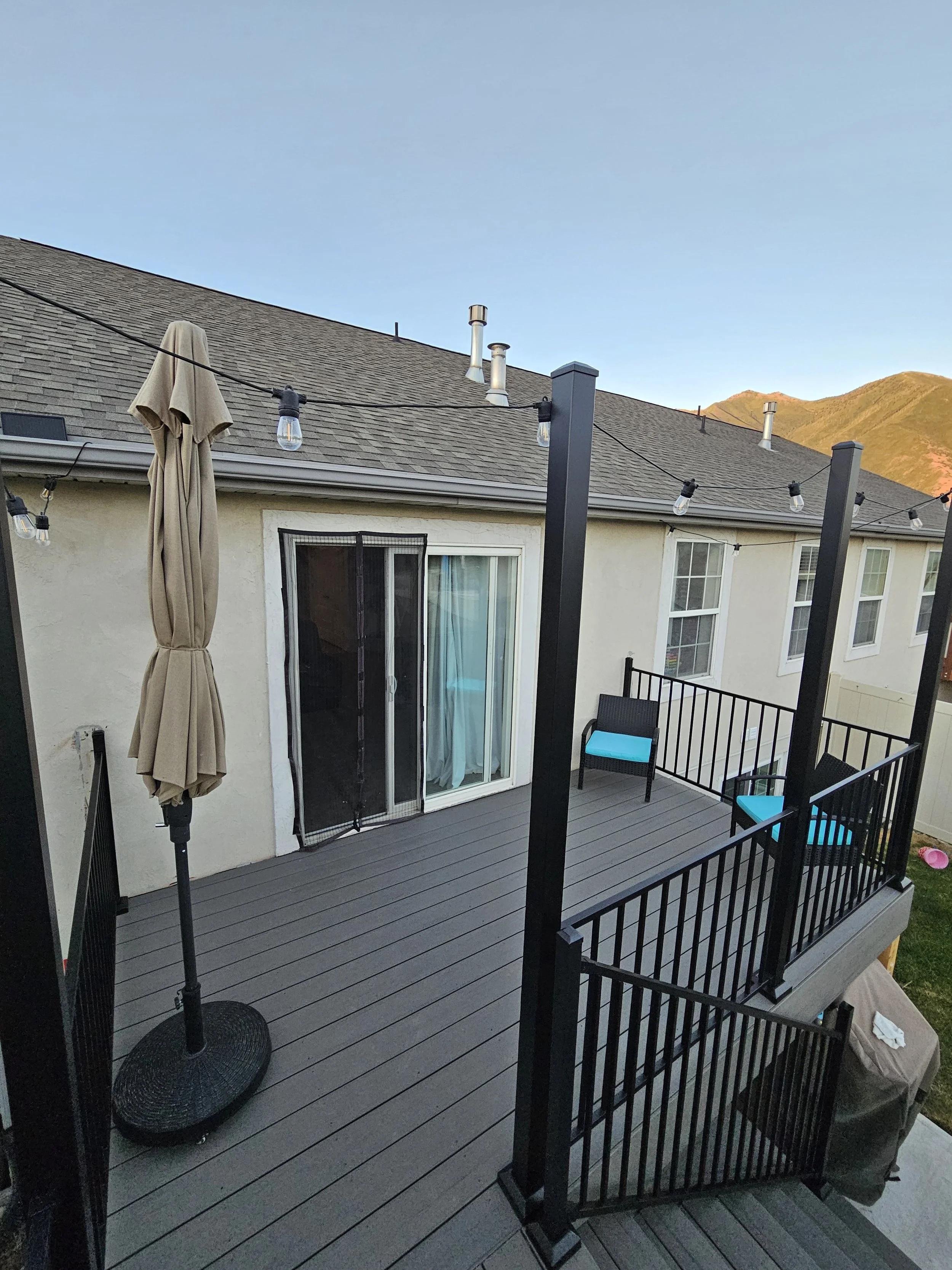 A backyard deck with black railings, patio furniture with blue cushions, string lights, a closed beige umbrella, and a sliding glass door leading into a house. Mountains are visible in the background.