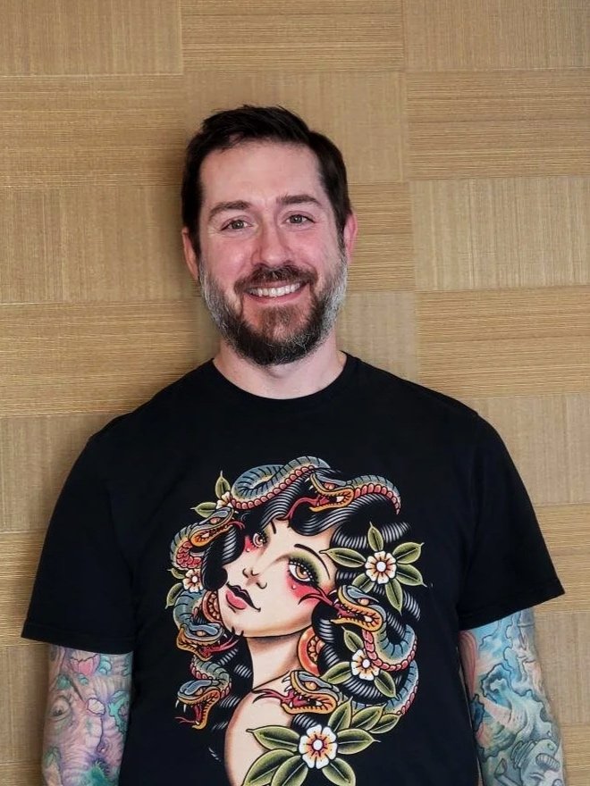 A man with a beard and tattoos on both arms, smiling, standing against a wooden wall. He is wearing a black T-shirt with a colorful illustration of a woman with dark hair, surrounded by flowers and snakes.