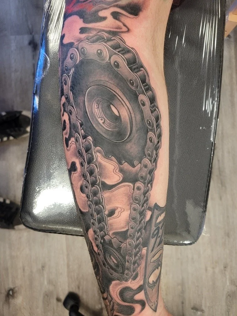 Black and gray tattoo of a motorcycle chain and sprocket on a person's arm with some background shading.