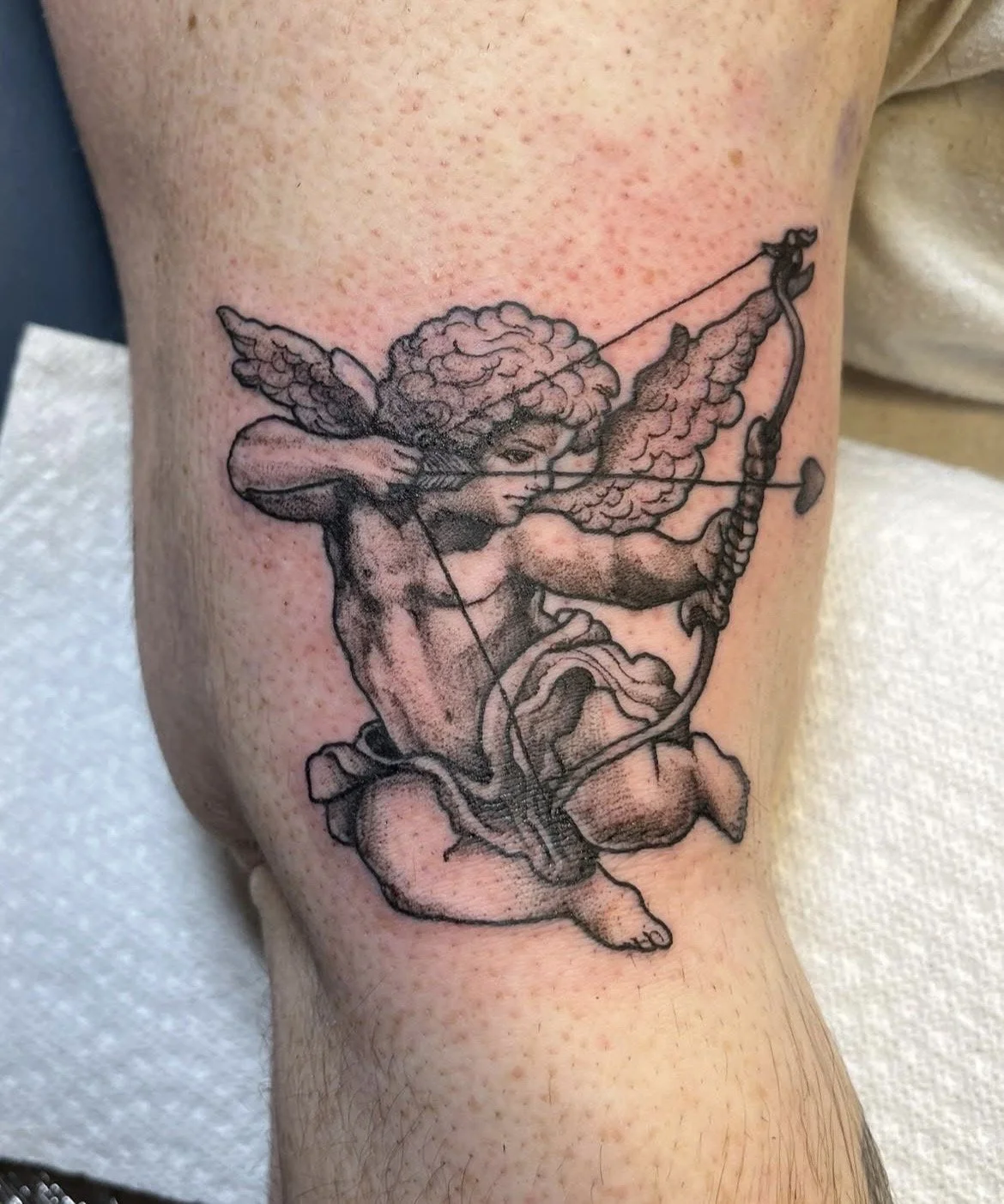 Black and gray tattoo of Cupid with curly hair, wings, and bow, aiming an arrow while kneeling.