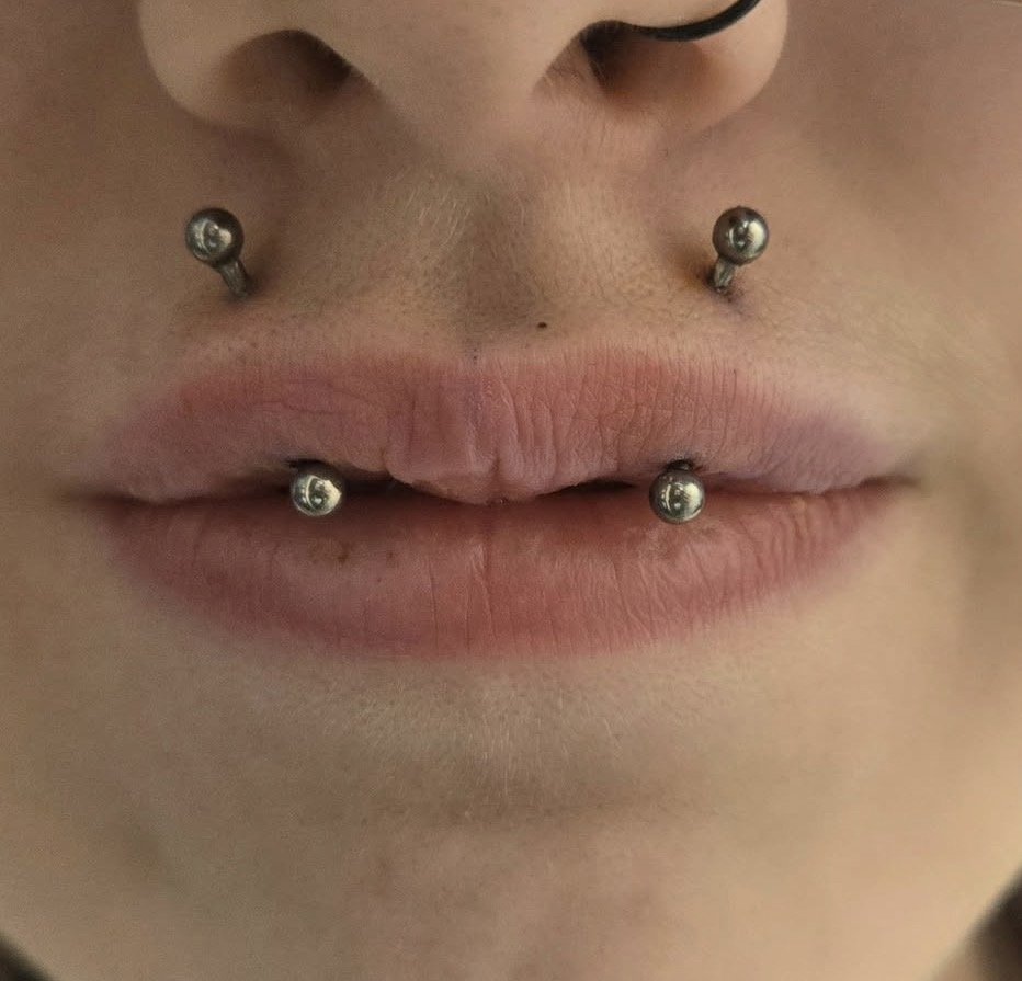 Close-up of a person's lips with multiple facial piercings, including two on the upper lip and two on the lower lip.