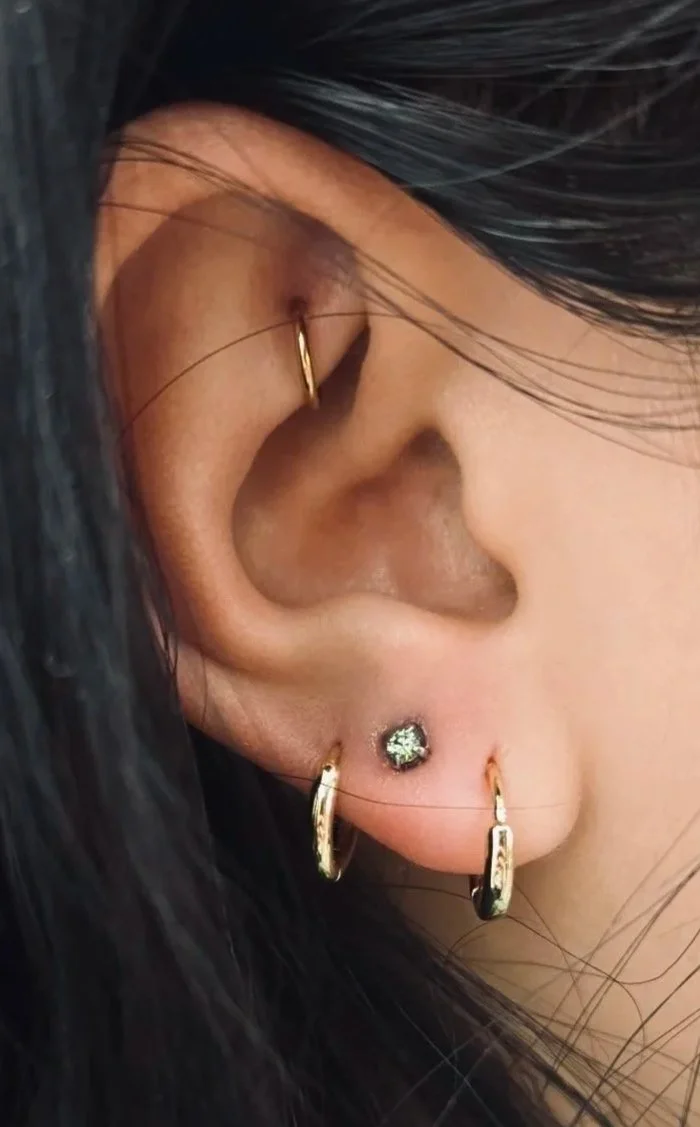 Close-up of a person's ear with multiple earrings, including gold huggie hoops, a small diamond stud, and a tiny hoop with a dangling charm, with black hair partially covering the ear.