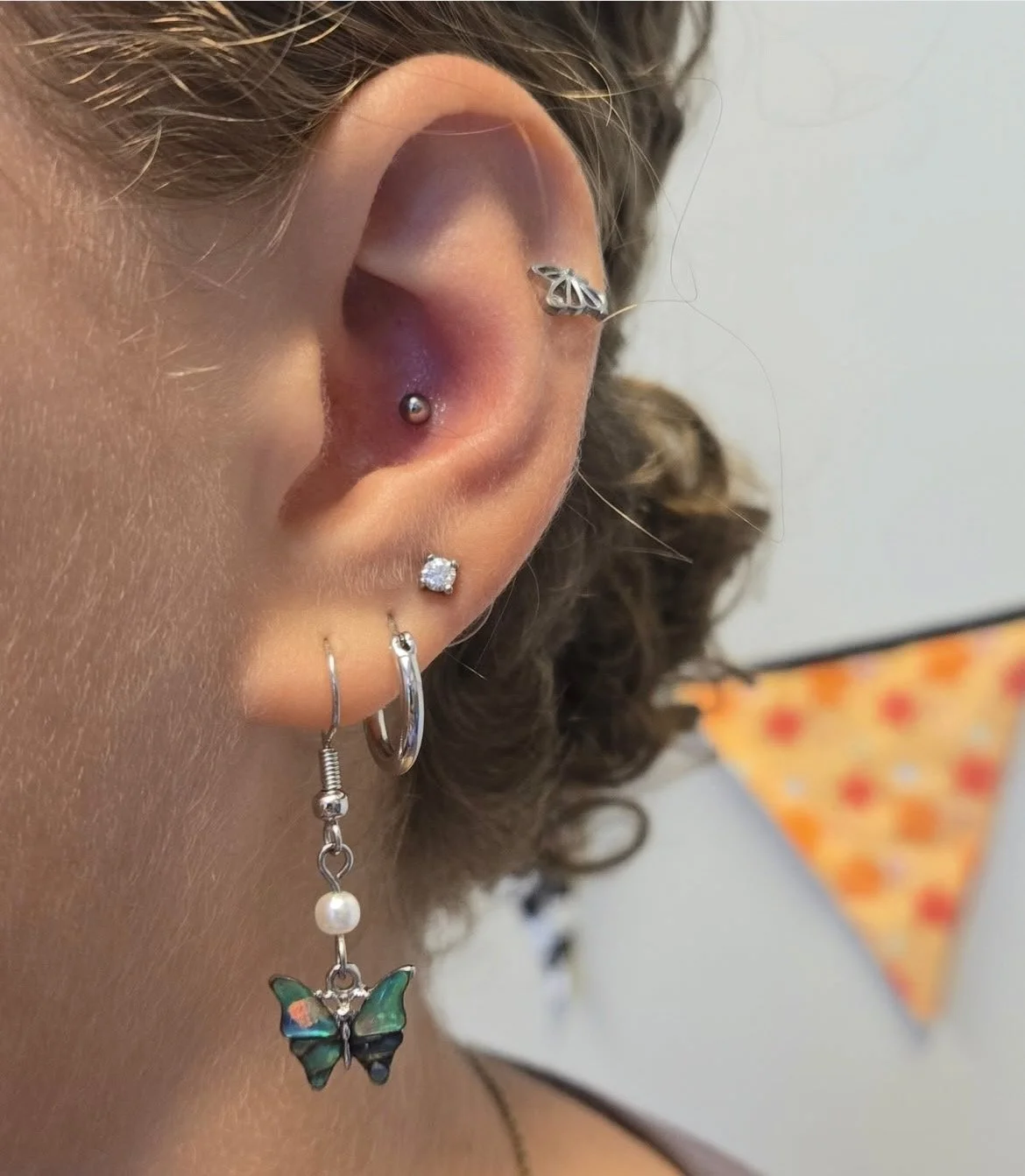 Close-up of a woman's ear with multiple earrings, including a silver butterfly cartilage ring, a helix piercing with a silver leaf-shaped earring, a dimple piercing with a silver ball, a stud earring with a clear gemstone, a small hoop earring, and a
