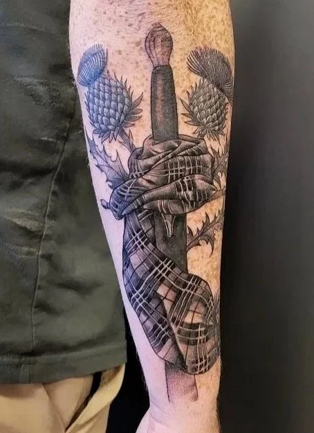 A tattoo on a person's arm featuring a dagger wrapped in a tartan cloth, surrounded by two pineapples with detailed leaves.