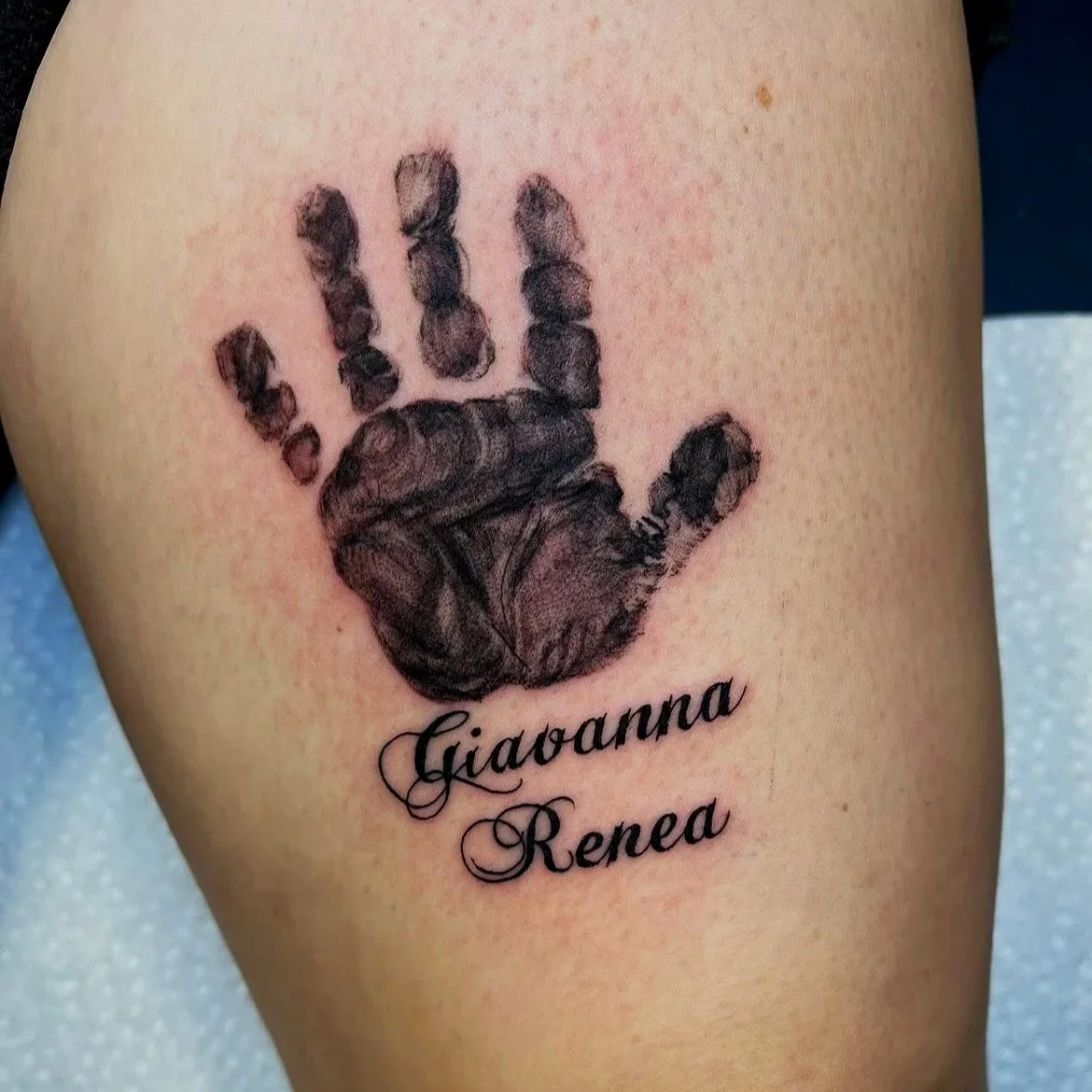 Tattoo of a realistic black ink baby handprint with the name Giaovanna Renea written below in cursive.