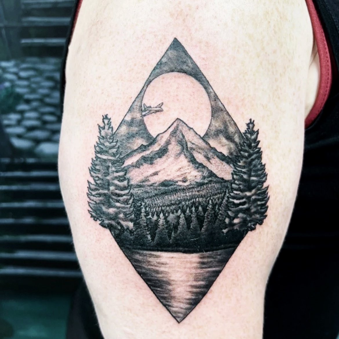 Black ink tattoo featuring mountains, pine trees, a lake, and a moon or sun with an airplane flying near it, within a diamond-shaped frame.