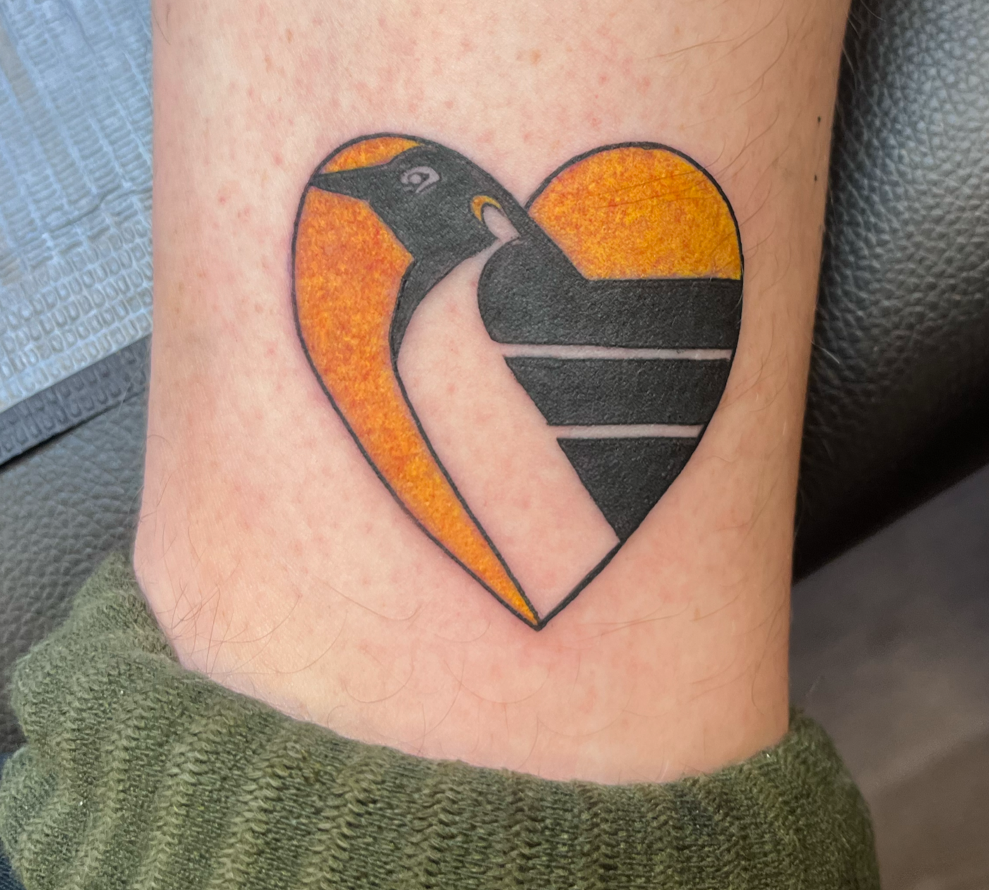 Tattoo of a penguin inside a heart shape with orange and black colors.