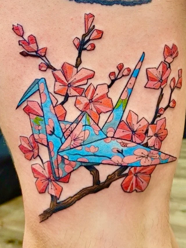 Colorful tattoo of a blue origami crane perched on a cherry blossom branch.