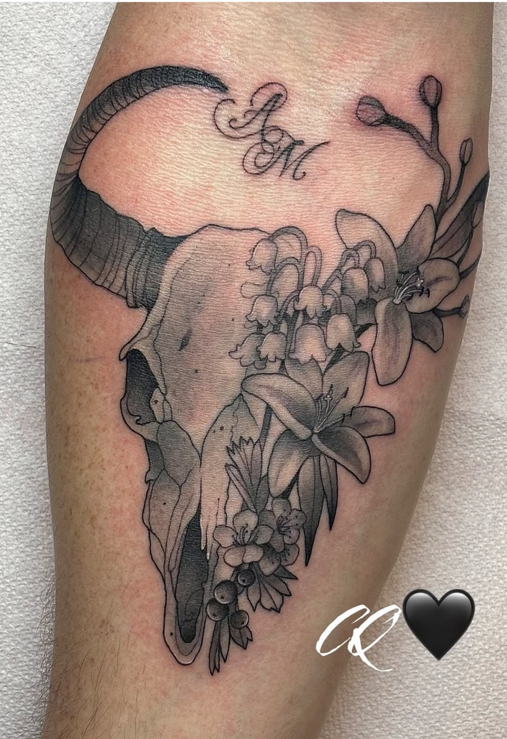 Black and gray tattoo of a bison skull with floral elements, featuring a detailed horn, flowers, and leaves, with the initials 'A.M.' above. There is a small 'CQ' and a black heart symbol near the bottom.