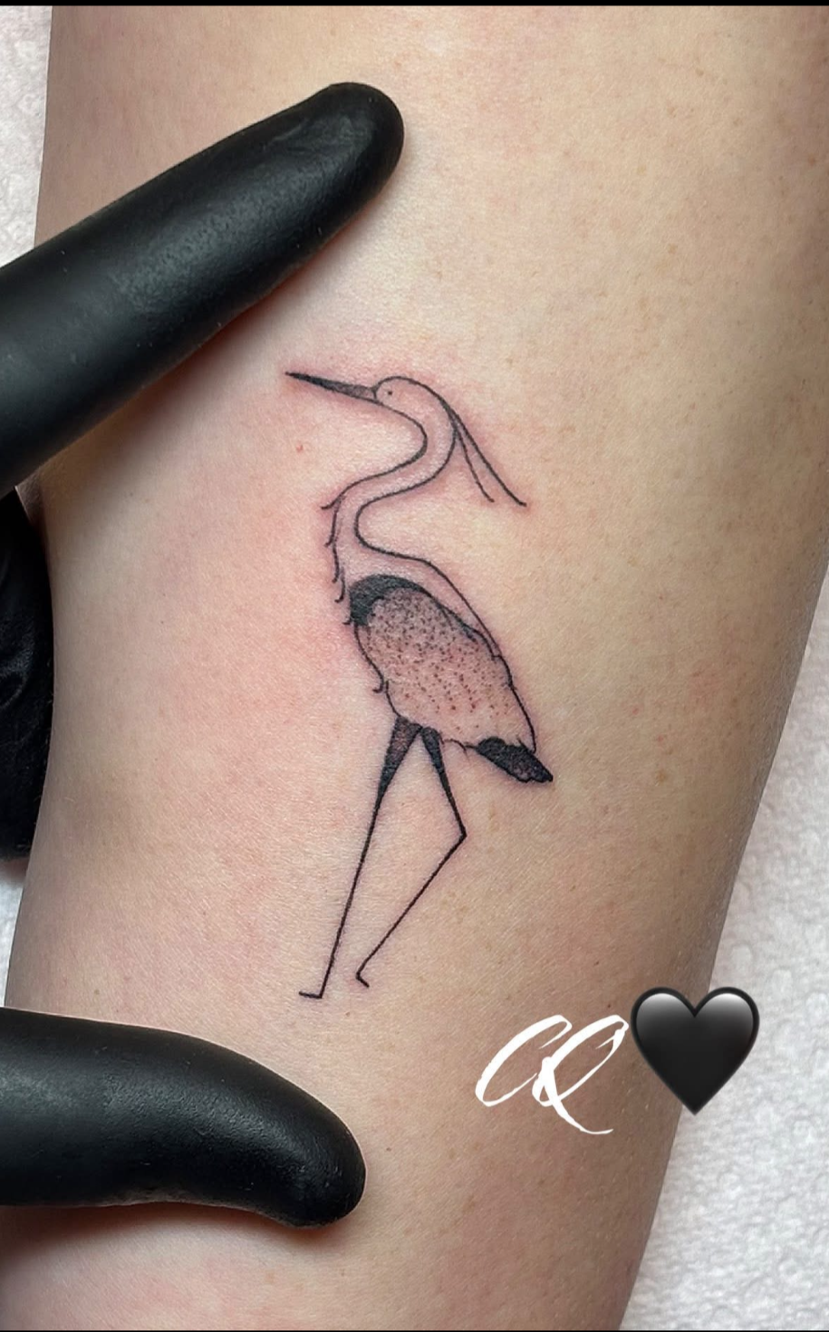 A tattoo of a minimalistic drawing of a flamingo with a long neck, detailed feathers, and thin legs on skin, with a black heart and initials 'CQ' in white nearby.