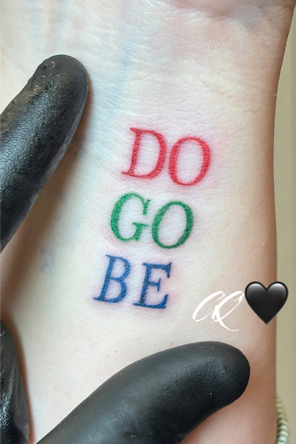 Tattoo with the words 'DO GO BE' in red, green, and blue, with a black heart and white decorative initials underneath.