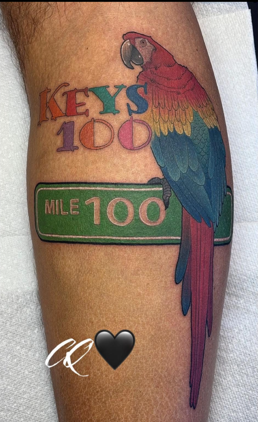 Colorful tattoo of a parrot sitting on a green mile marker sign that reads 'MILE 100'. Above the parrot, the words 'KEYS 100' are written in multicolored letters. There is also a black heart and a white signature or initials below the sign.