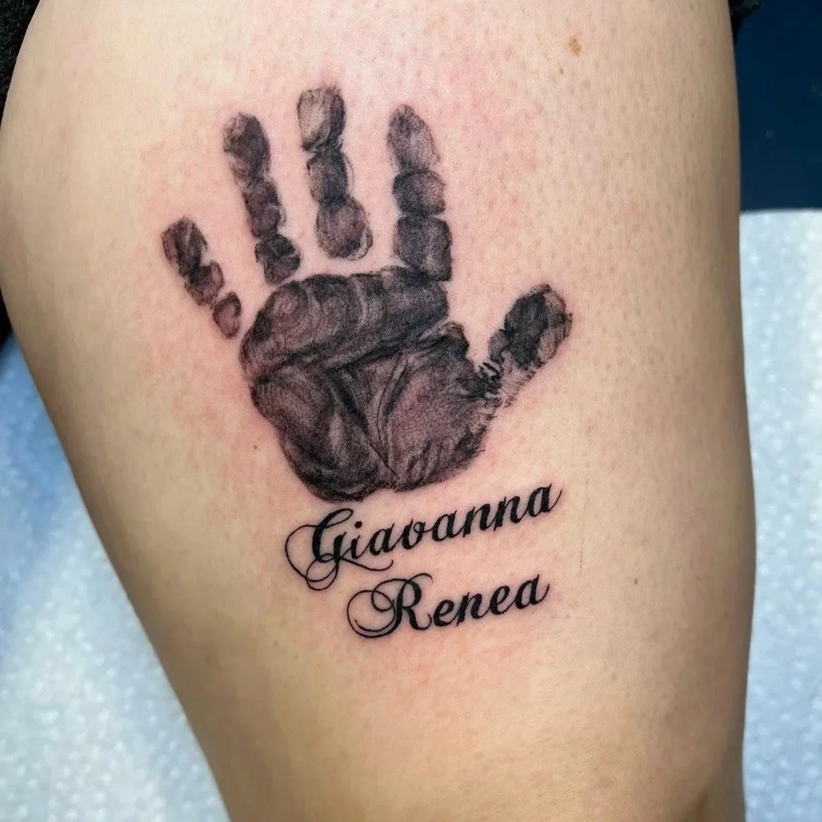 A tattoo of a realistic black ink handprint with the names Giaovana Renea written in elegant cursive below.