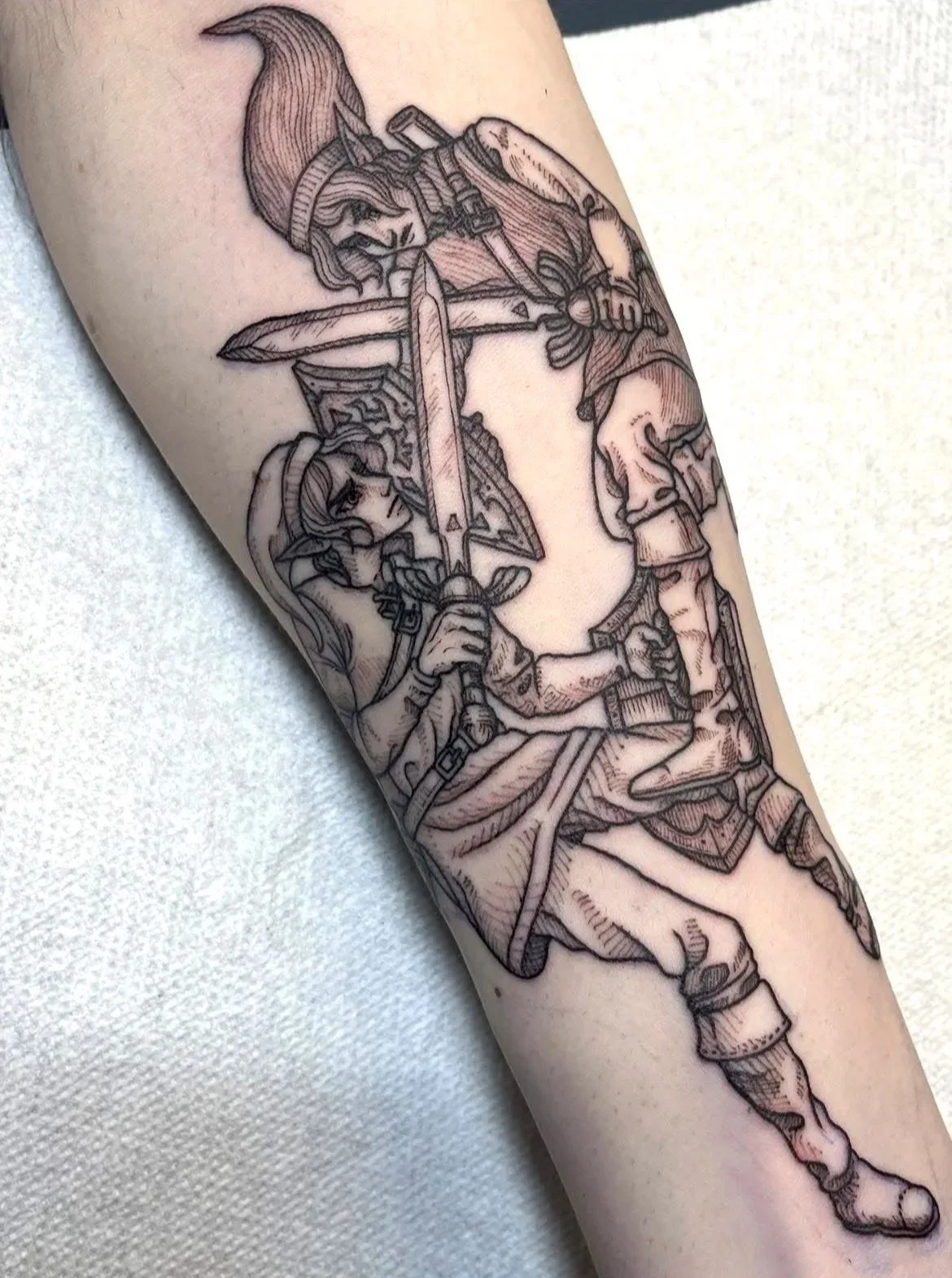Black and gray tattoo of two characters dressed as fantasy warriors with swords and shields, sitting on a surface, on a person's arm.