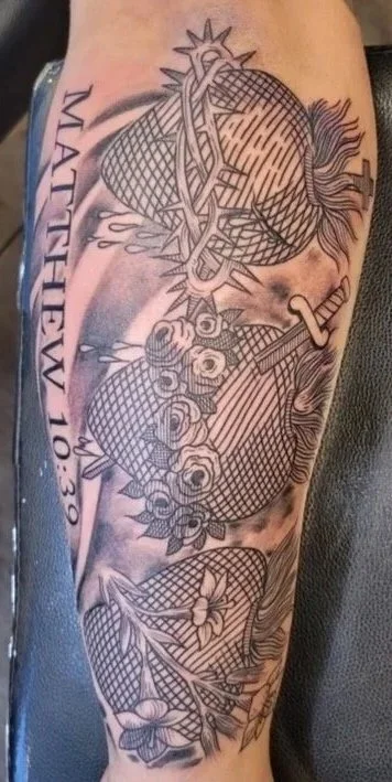 Tattoo of a large heart with a chain wrapped around it, surrounded by roses, a dagger, and the words "MATTHEW 10:30" on a person's arm.