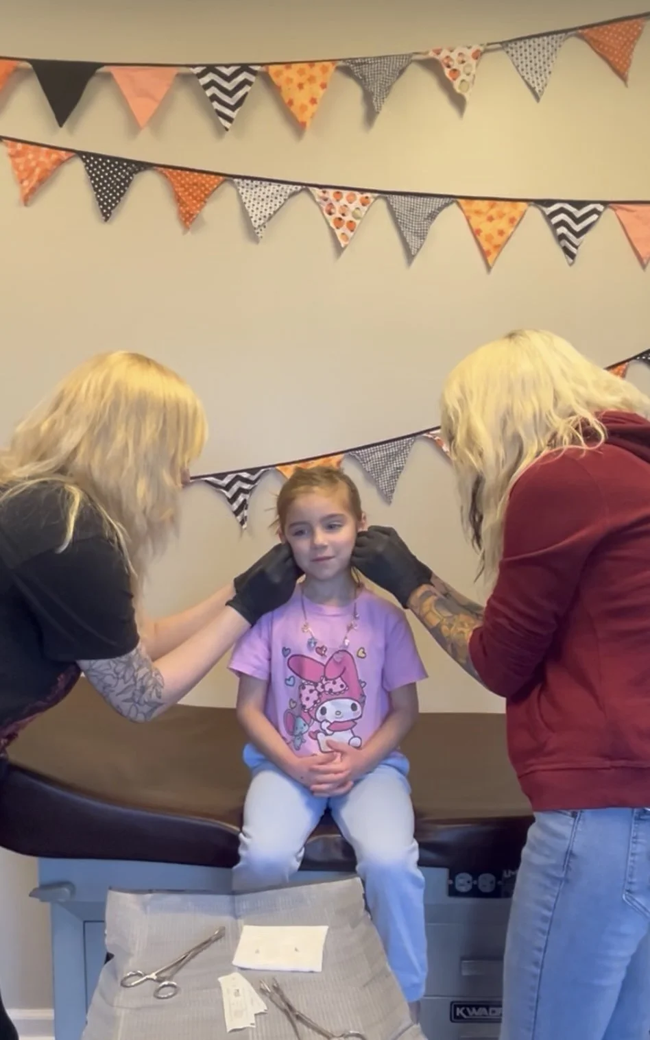 A young girl sitting on an examination table while two professionals, possibly medical staff, prepare her for a procedure. The girl is wearing a pink graphic T-shirt and white pants. One professional is adjusting the girl's ear, and the other is assi