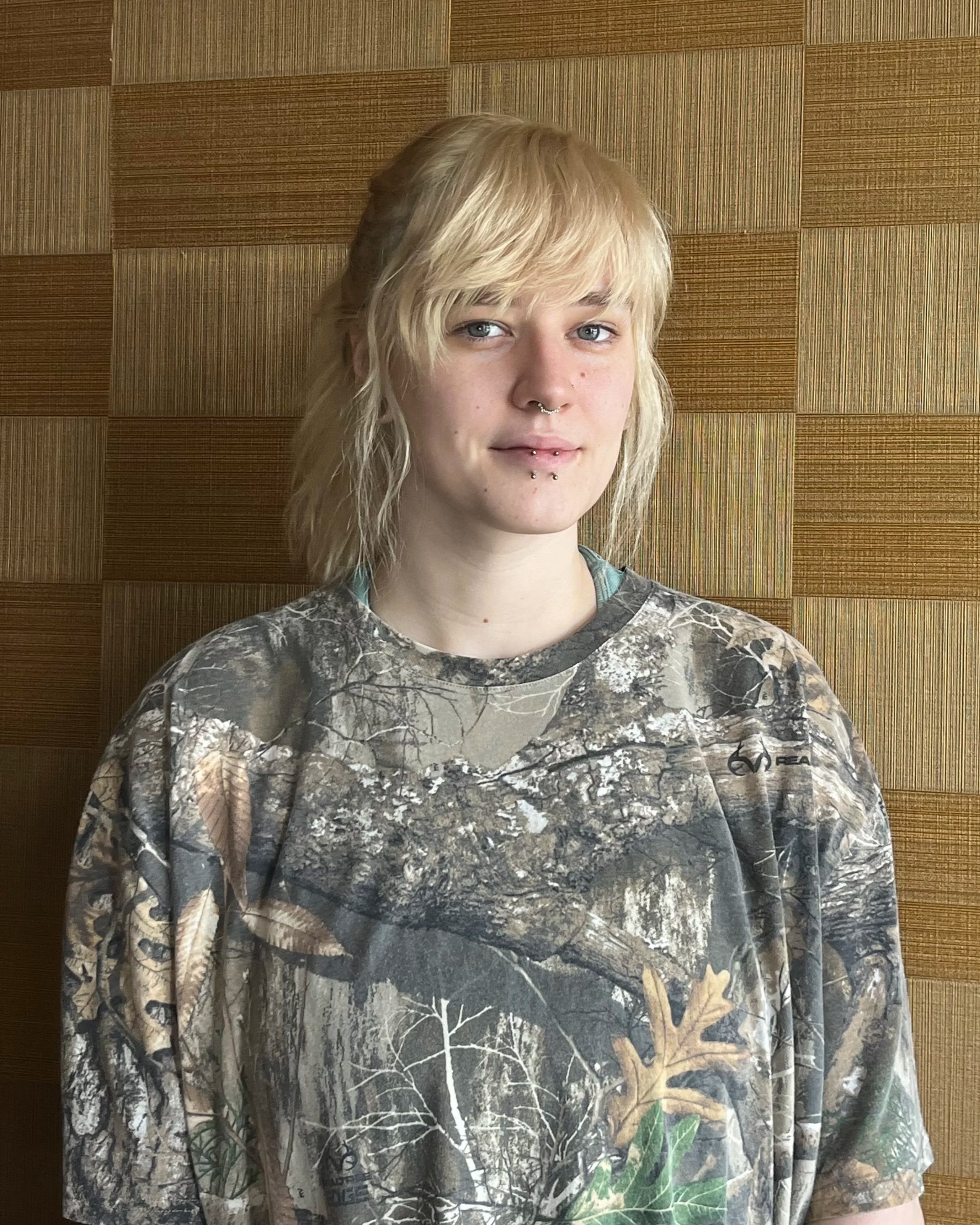 A young woman with blonde, shoulder-length hair and a septum piercing, standing in front of a wooden paneled wall, wearing a camouflage sweatshirt.