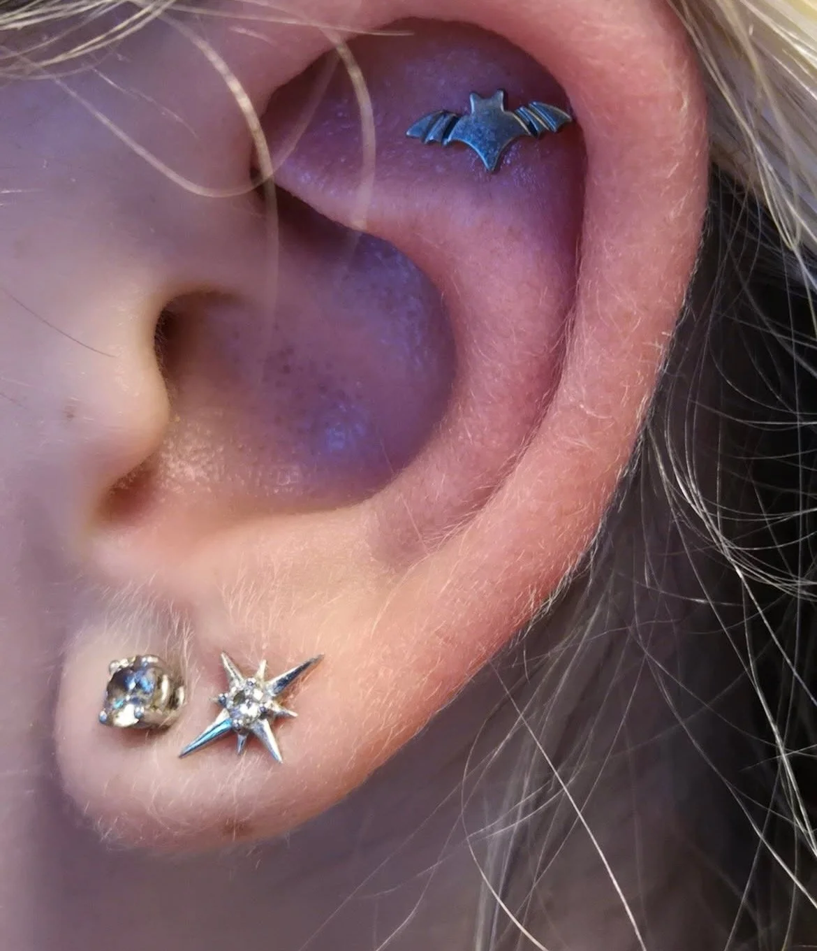 Close-up of a person's ear with three piercings. The upper piercing is a bat-shaped jewelry in the helix, and the lower lobe piercings are a heart-shaped stud and a star-shaped stud with small gemstones.