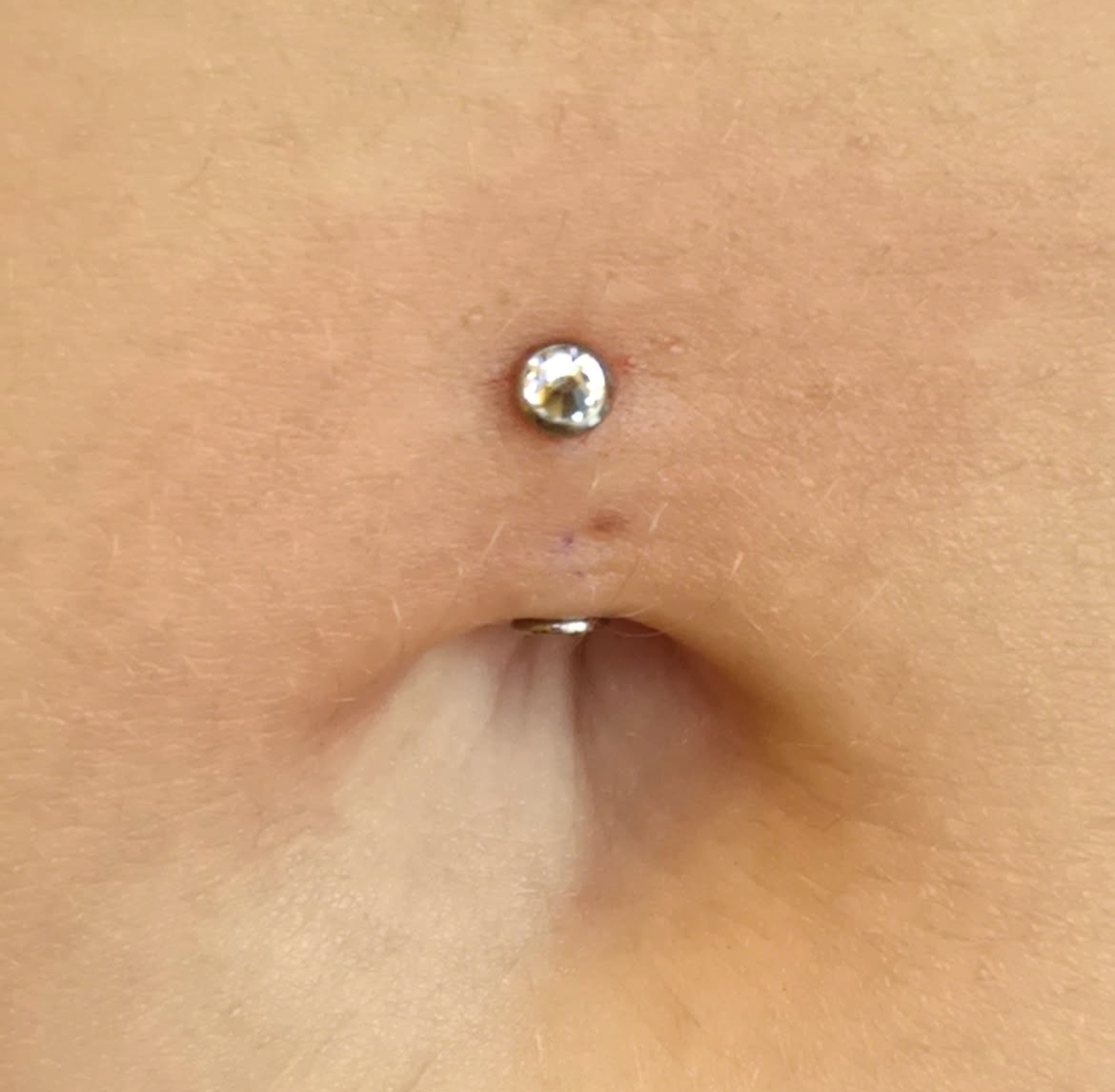 Close-up of a person's belly button with a small silver piercing that has a clear gemstone, and a second piercing below it.