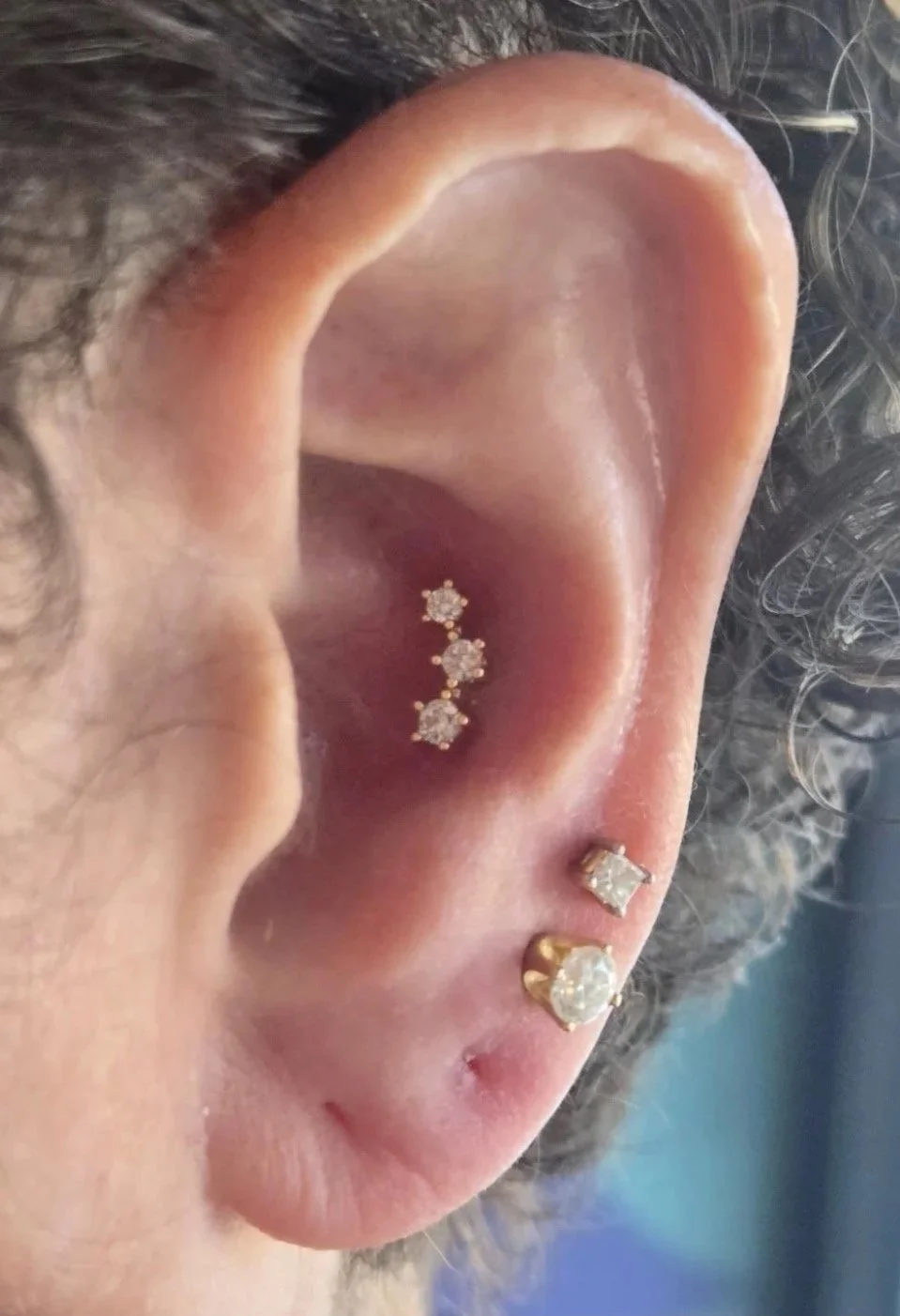 Close-up of a person's ear with multiple earrings. The earrings include a row of three small diamond studs along the upper cartilage, a medium-sized diamond stud in the middle, and a larger diamond stud with a gold backing at the bottom.