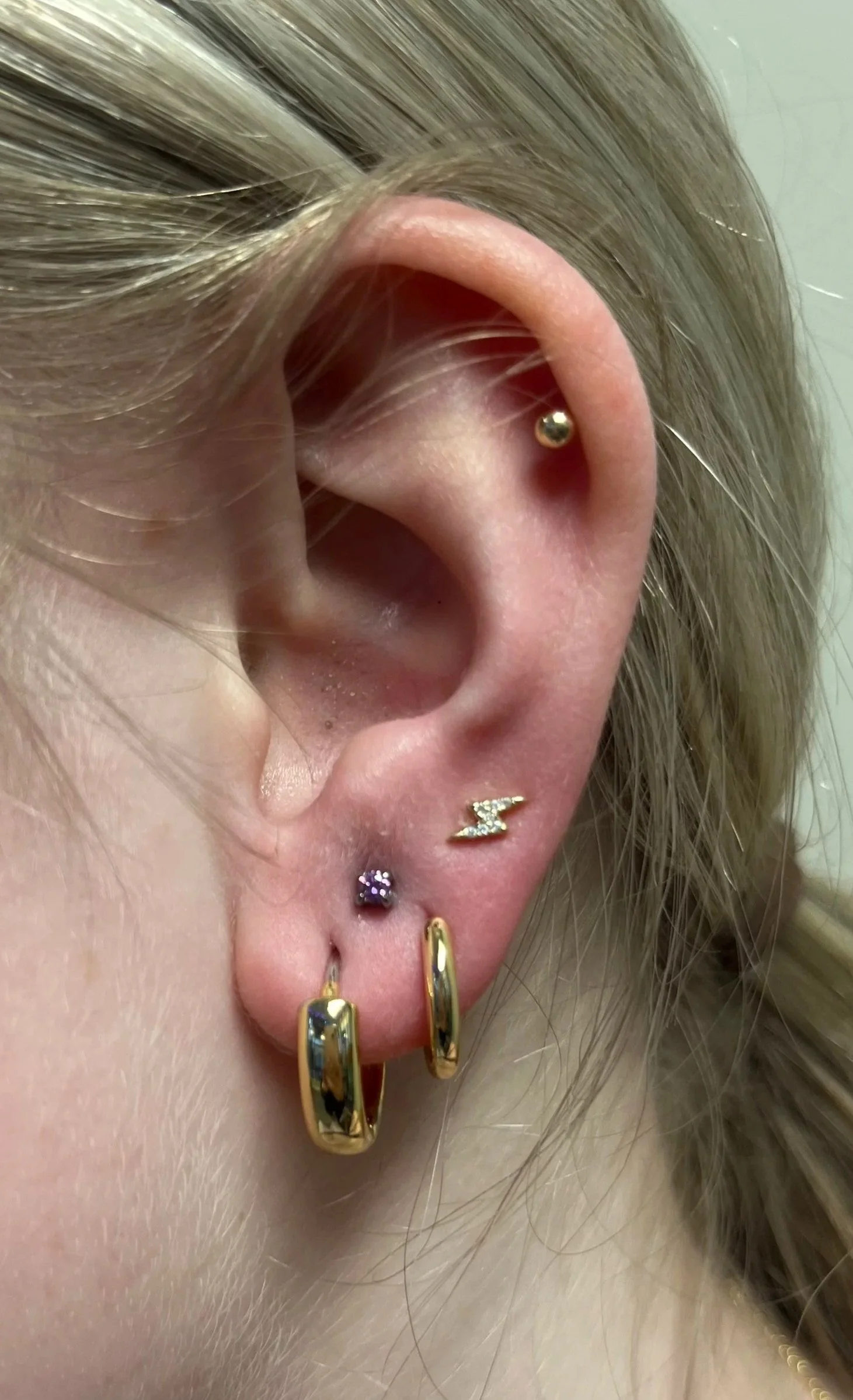 Close-up of a person's ear with multiple earrings: a gold ball stud in the upper cartilage, a small purple gemstone stud in the lobe, a lightning bolt-shaped gold earring, and a plain gold hoop earring.