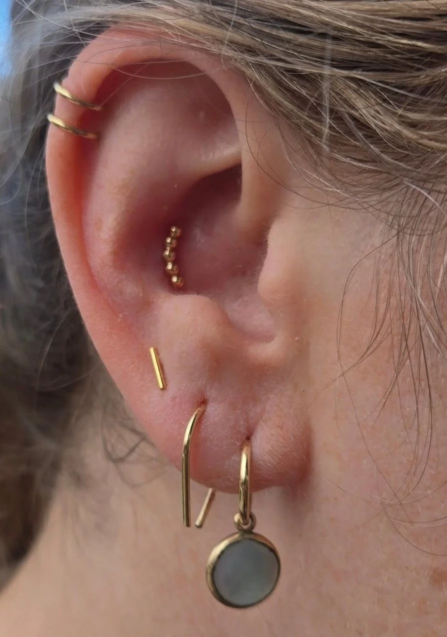 Close-up of a person's ear decorated with multiple gold earrings, including a small hoop with beads, a skinny bar, and a dangling earring with a round gray stone.