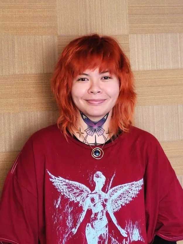 Young woman with short, wavy red hair smiling, wearing a red graphic t-shirt and a black choker with a circular pendant, standing against a light wood-paneled background.