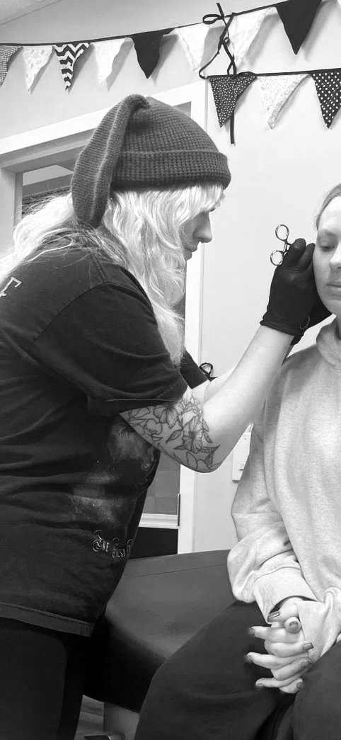A woman with long hair and a beanie hat is tattooing a young woman, who is sitting with her hands clasped. The tattoo artist has tattoos on her arm and is wearing gloves, working on the young woman's eyebrow or face. Decorated banners hang on the wall in the background.