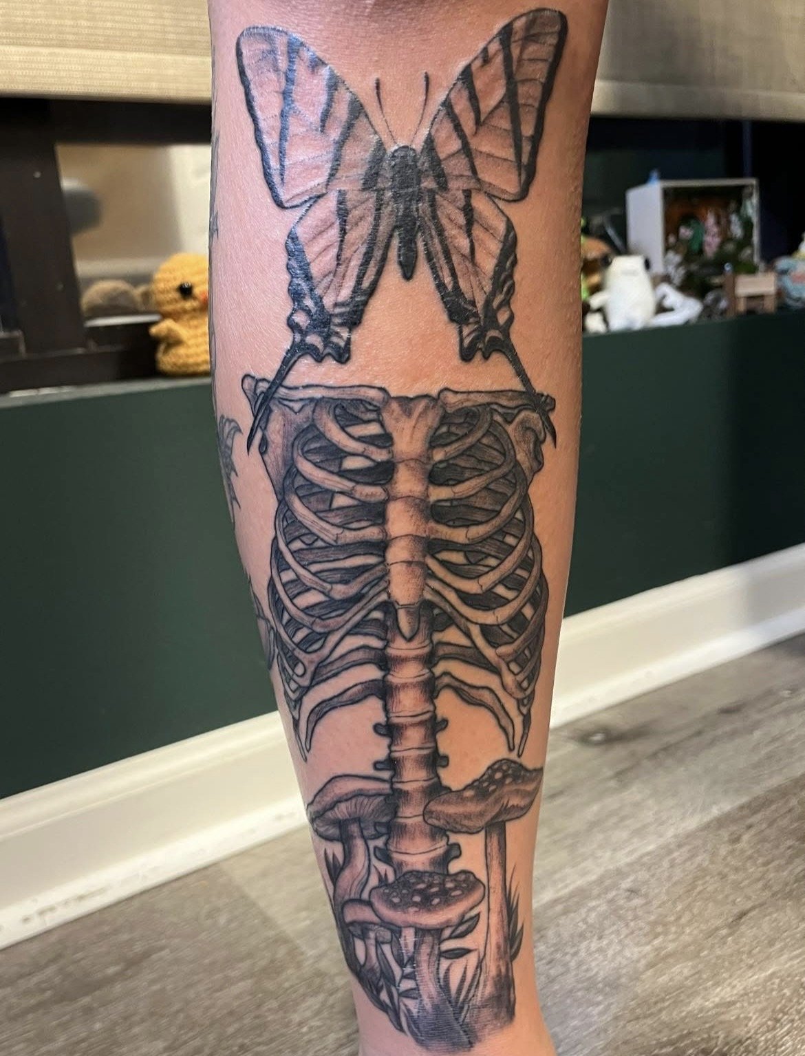 A tattoo on a person's lower leg showing a butterfly atop a skeleton with mushrooms growing around the bones.