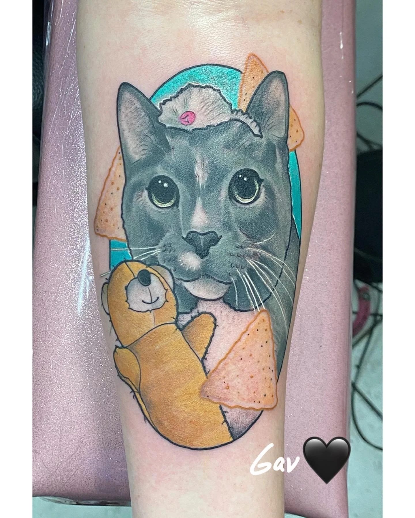 Colored tattoo of a realistic cat's face with large, shiny eyes, surrounded by decorative elements including a teddy bear, tortilla chips, and a teal circle, with the name 'Gav' and a black heart.
