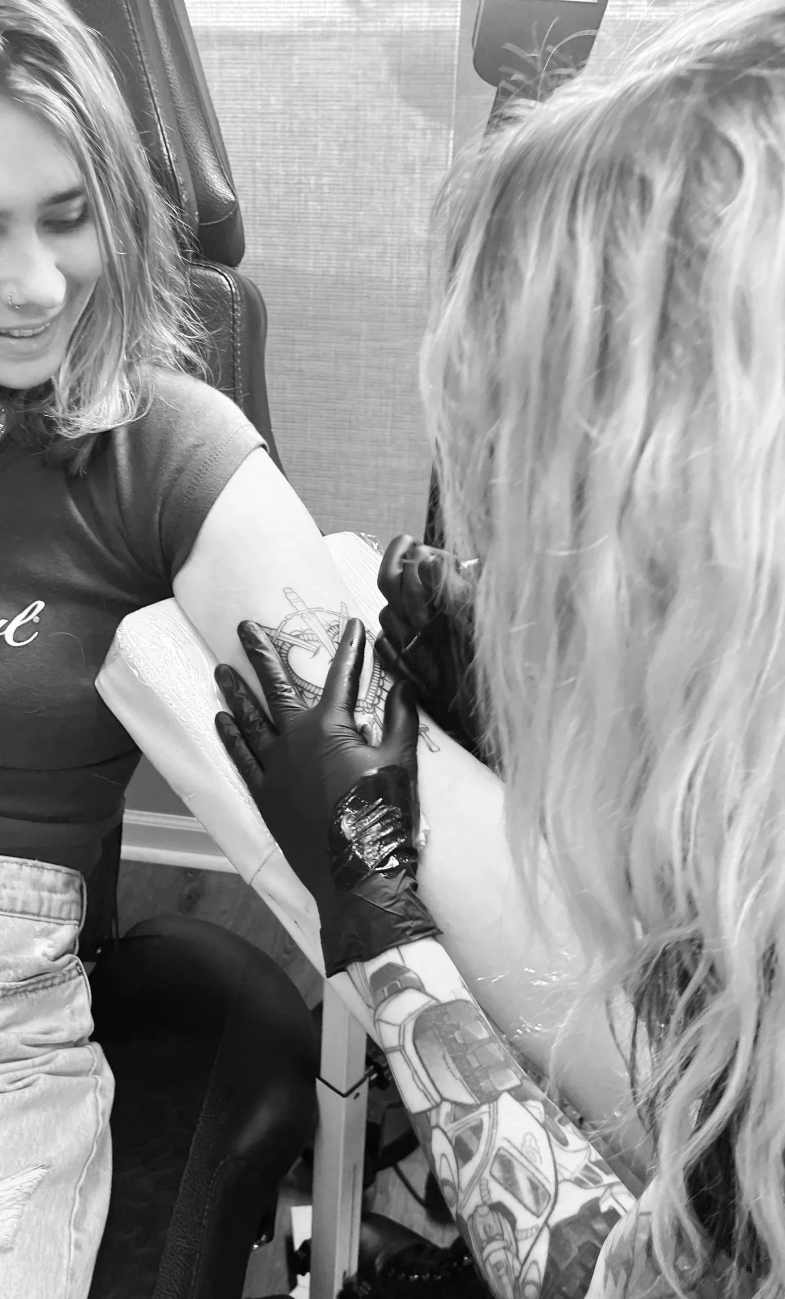 A person getting a tattoo on their upper arm by a tattoo artist. The person receiving the tattoo is smiling, and the artist is wearing gloves while working on the tattoo design.